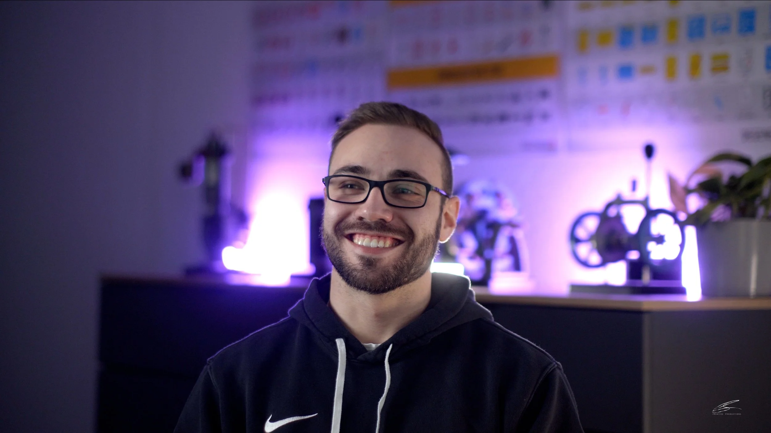 Ein lachender Mann mit Brille vor einem beleuchteten Hintergrund mit modernen Dekorationen und einem Pflanzen.