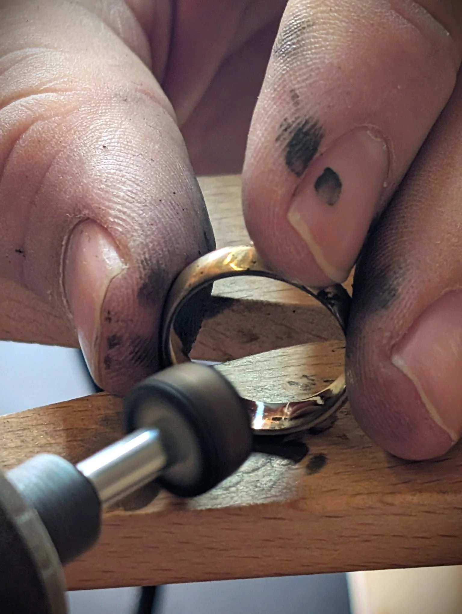 Close-up of a person's fingers with black smudges holding a ring and working on it with a rotary tool on a wooden surface.