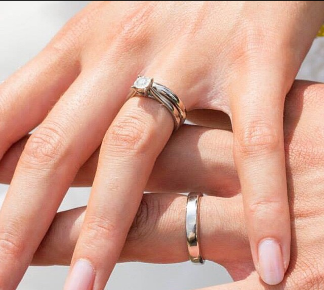 Close-up of two hands with wedding rings; one hand has a diamond engagement ring and a wedding band, the other hand has a plain wedding band, both intertwined.