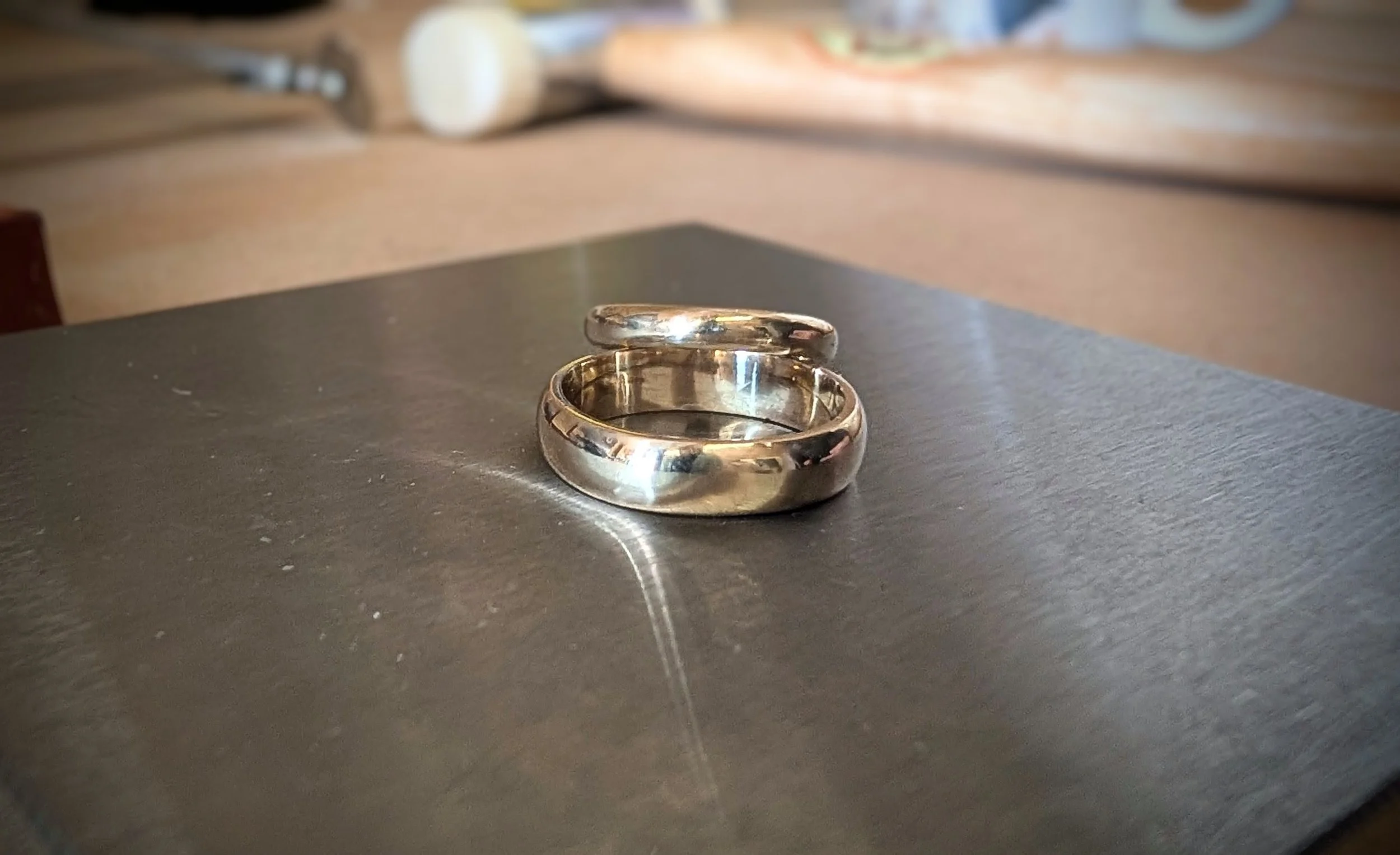 A pair of gold rings, one with a thicker band, placed on a dark surface with a blurred background.