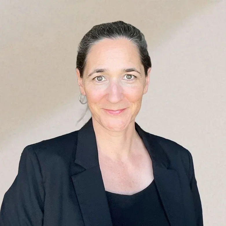 Eine Frau mit dunklem Haar, die einen schwarzen Blazer trägt, vor einem hellen Hintergrund, lächelnd in die Kamera schauend.