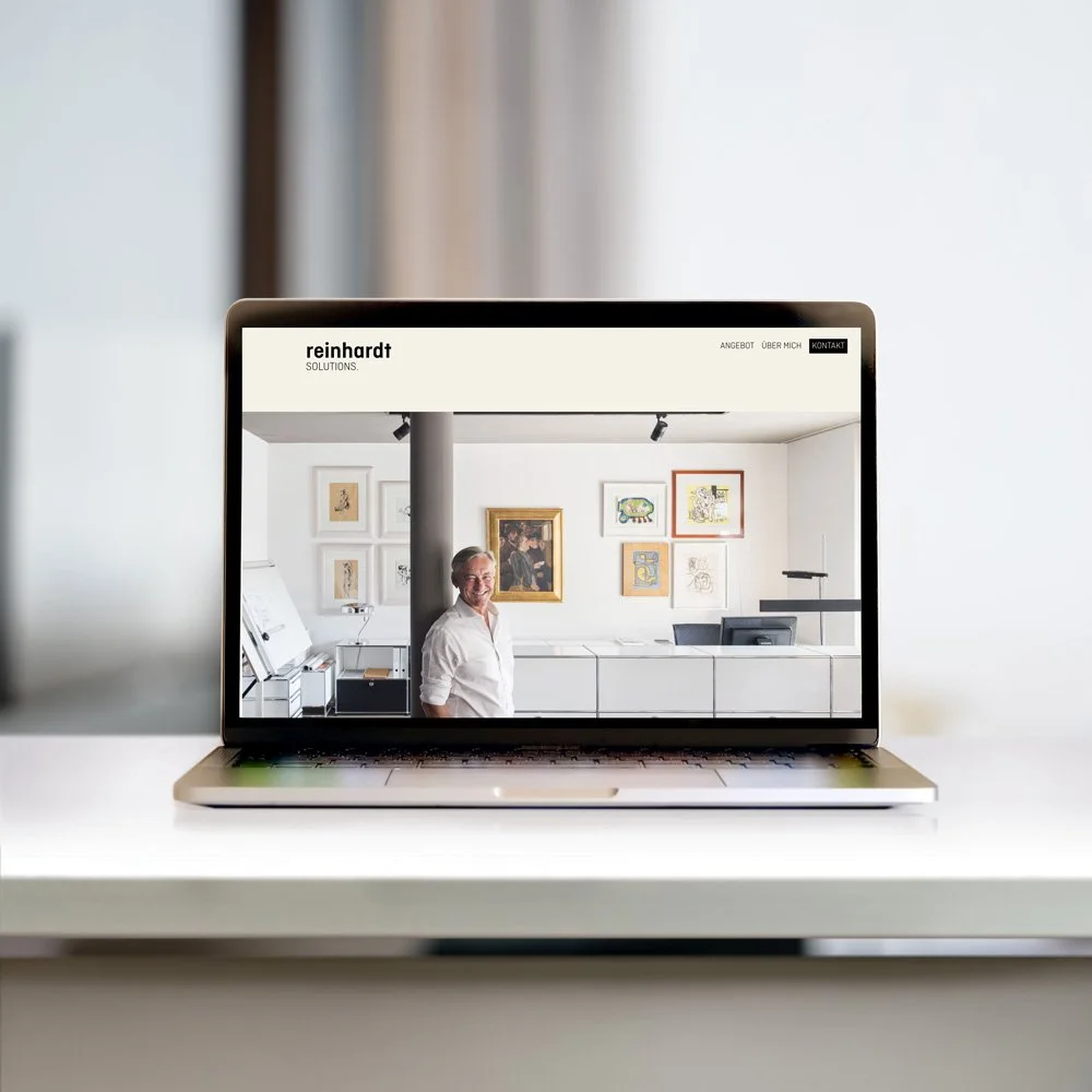 Ein Laptop zeigt eine Webseite mit einem Bild von einem Mann in einem Kunstgalerie oder Büro, der vor einer Wand mit mehreren Kunstwerken steht.