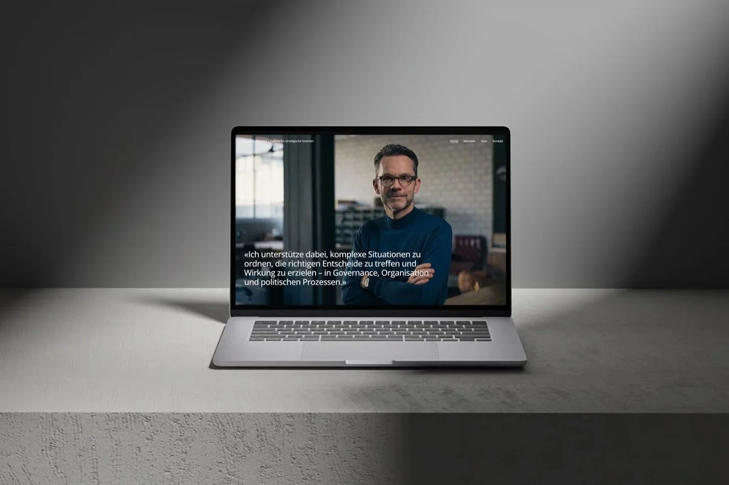 Laptop auf einem Tisch mit einer Webseite, showing einen Mann mit Brille und dunklem Pullover, der vor einem Bücherregal steht, mit deutschen Text auf dem Bildschirm.
