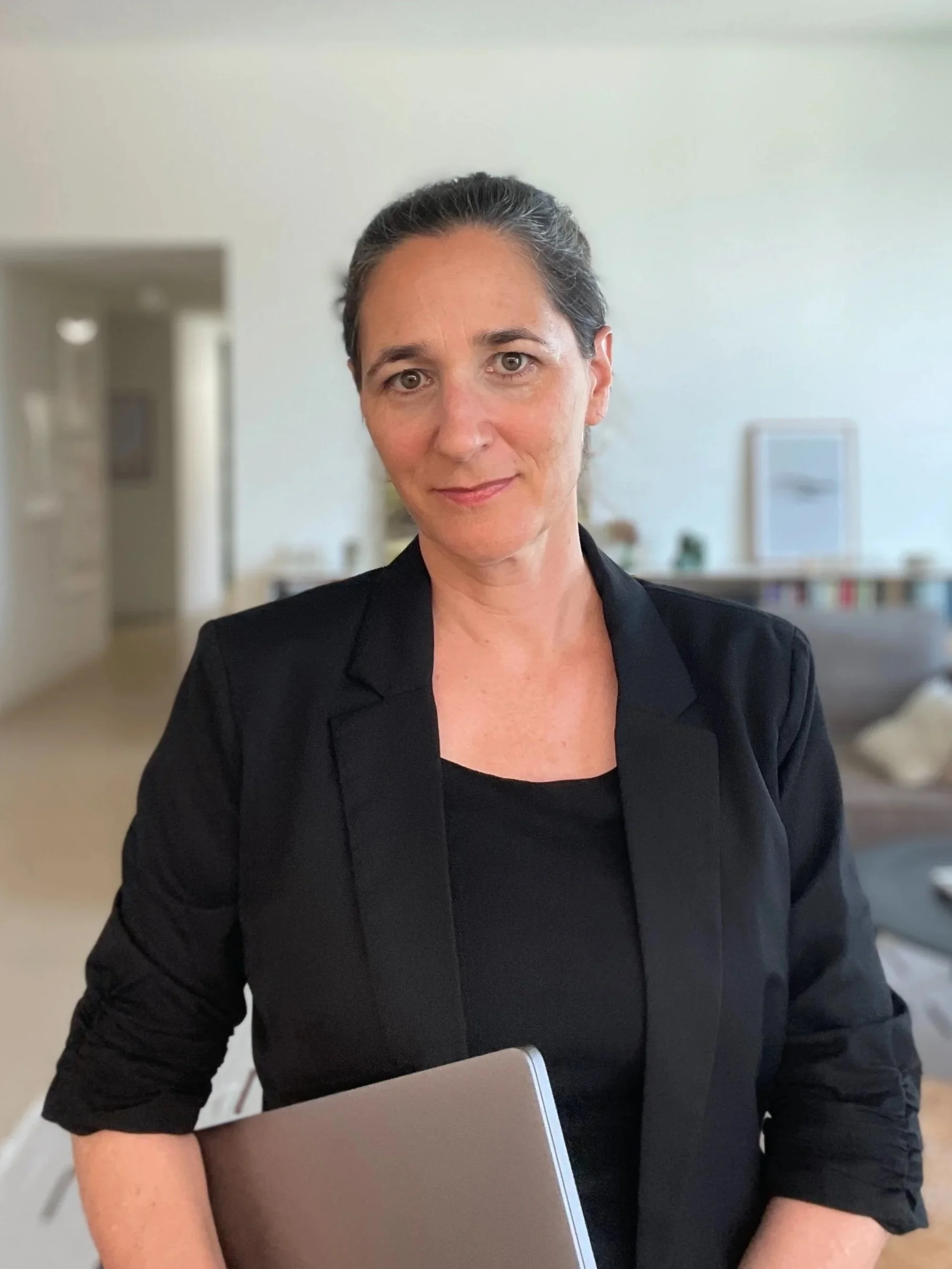 Eine Frau mit dunklen Haaren in einem schwarzen Blazer hält einen Laptop in den Armen, im Hintergrund ein helles Wohnzimmer.