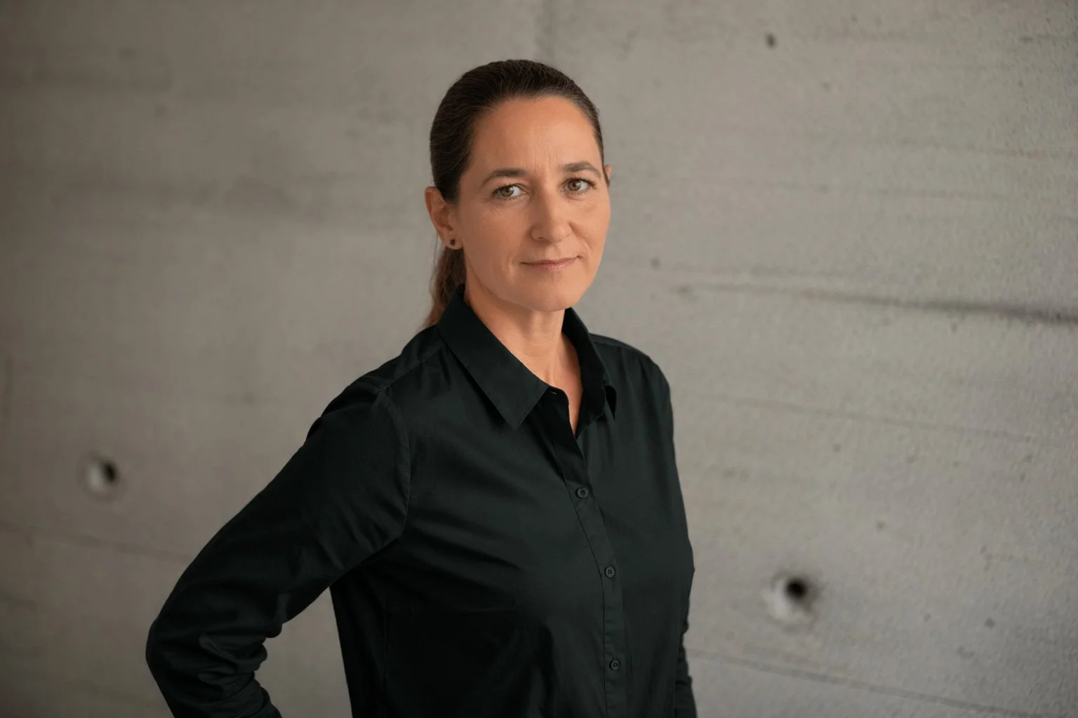 Eine Frau trägt eine schwarze Bluse und steht vor einer grauen Betonwand.