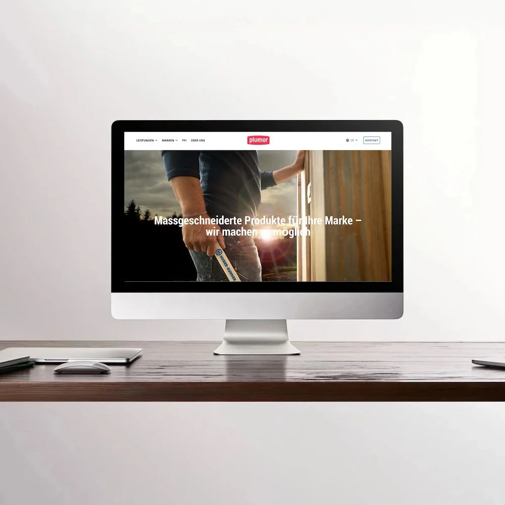 Ein Computerbildschirm zeigt eine Webseite der Firma Plumor mit dem Slogan 'Massgeschneiderte Produkte für Ihre Marke – wir machen möglich'. Der Bildschirm steht auf einem Holzschreibtisch mit einer Maus und einem Laptop. Der Hintergrund ist eine weiße Wand.