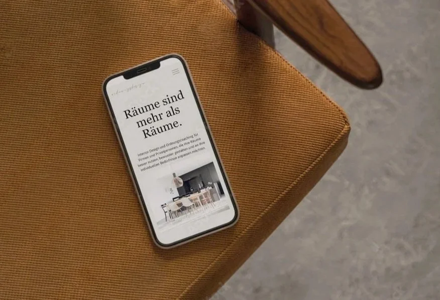 Ein Smartphone liegt auf einem braunen Sitzkissen. Auf dem Bildschirm ist ein Text in Deutsch zu sehen, der über Räume und Innenarchitektur spricht, begleitet von einem Bild eines modernen Esszimmers.