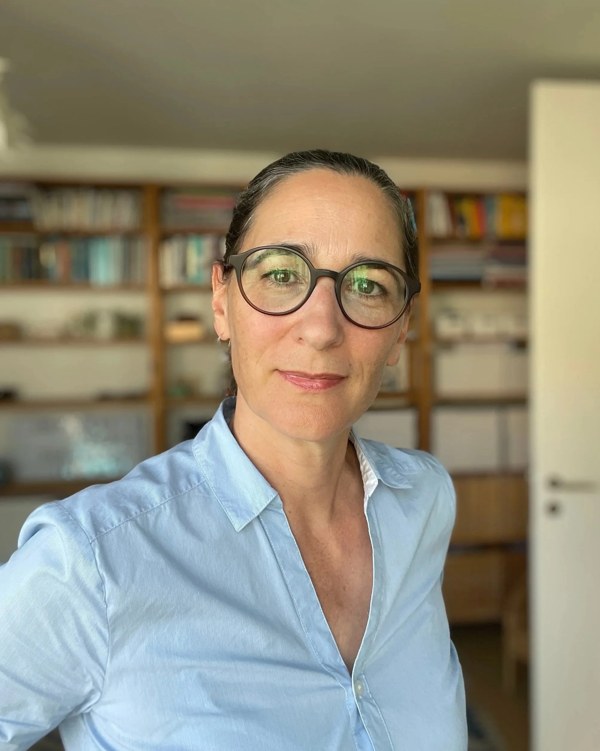 Porträt einer lächelnden Frau mit dunklen Haaren, trägt eine Brille und eine hellblaue Bluse, im Hintergrund ein Regal mit Büchern, Innenraum.