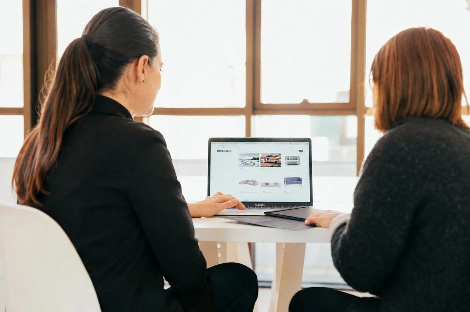 Zwei Frauen sitzen an einem weißen Tisch vor einem Fenster, schauen sich Produkte auf einem Laptop-Display an, das Möbel zeigt.