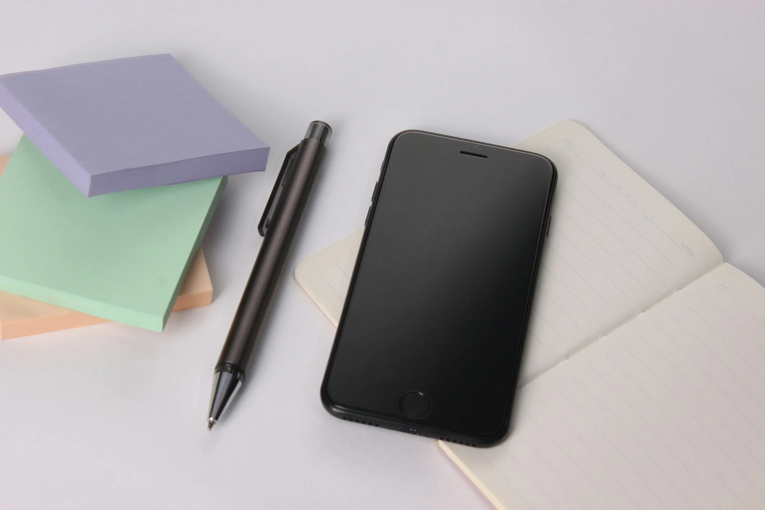 Black smartphone, silver pen, open notebook, and pastel sticky notes on white surface.