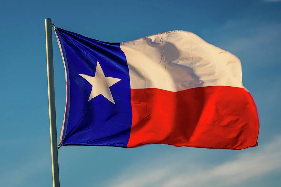 A Texas state flag flying outdoors against a cloudy sky.
