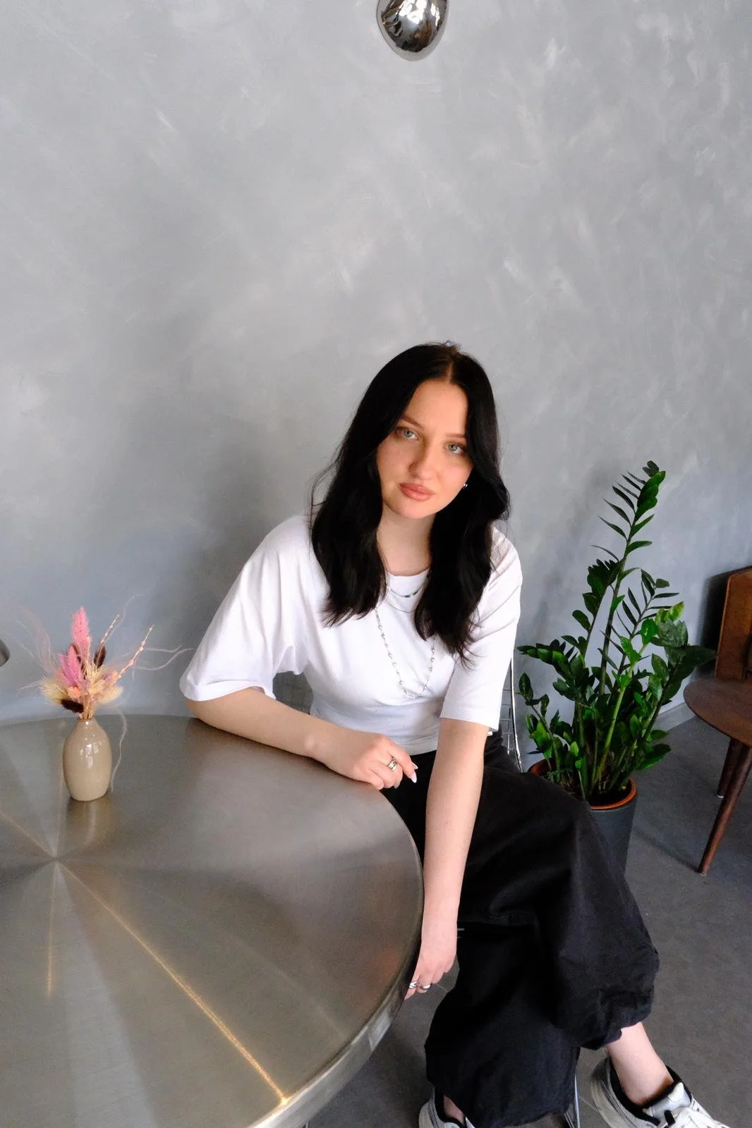 A young woman with black hair and light makeup sitting at a round metallic table in a modern, minimalistic room. She is wearing a white t-shirt, black pants, and sneakers, with a necklace and ring. On the table, there is a small beige vase with pink, beige, and brown dried flowers. Behind her, there's a large green plant in a pot and a wooden chair. The wall is grey and textured.