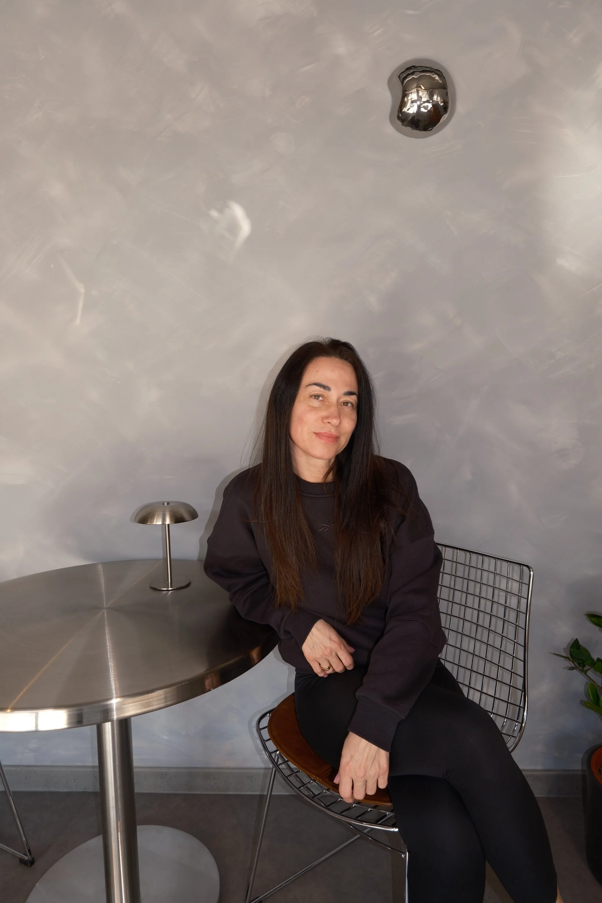 A woman with long dark hair, wearing a black sweatshirt and black pants, seated at a round metallic table with a small modern lamp. She is smiling gently and has her left arm resting on the table, with a wire chair behind her. The background includes a gray wall with a metallic balloon attached to the ceiling.