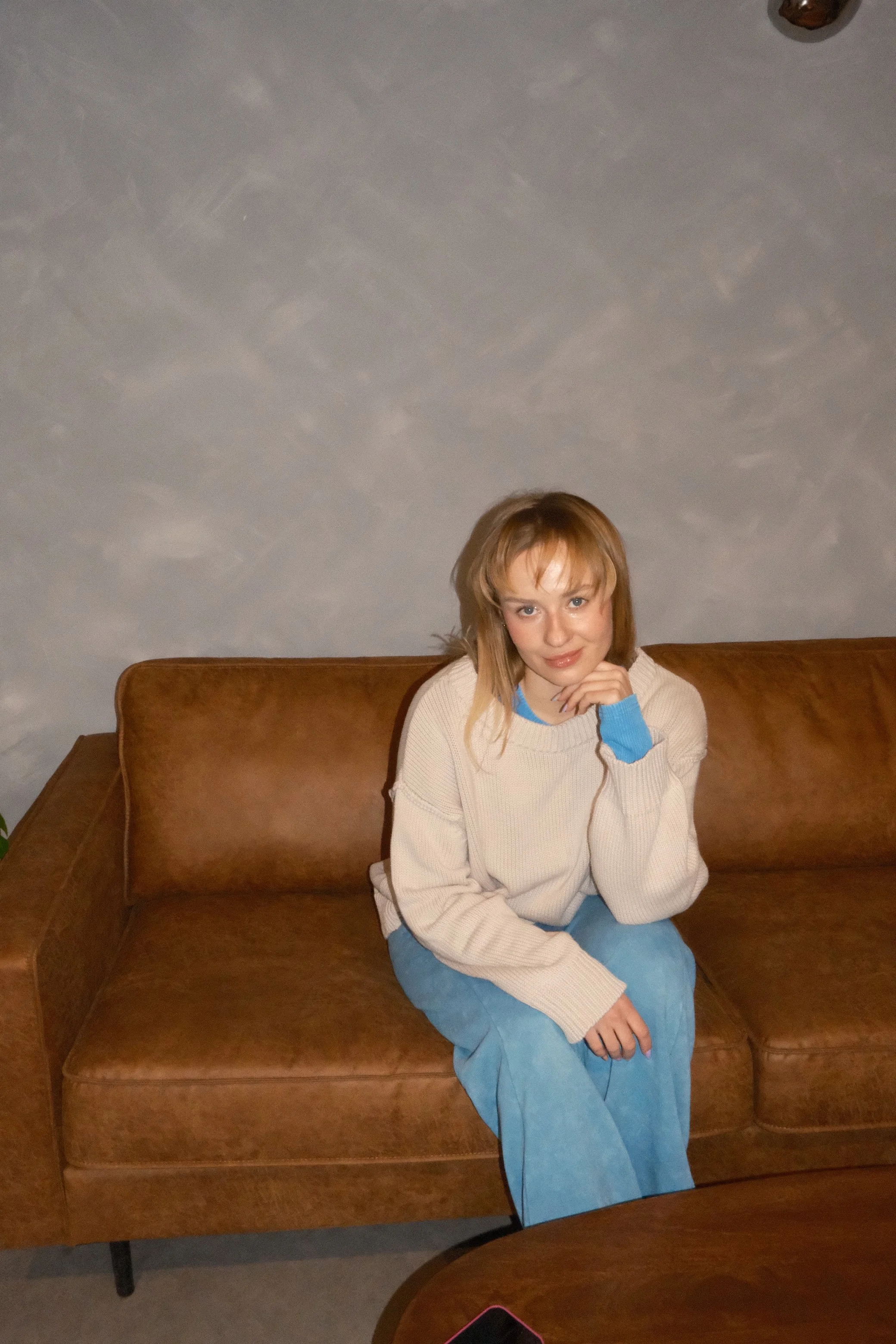 A young woman with light brown hair, wearing a cream sweater with blue accents and blue pants, is sitting on a brown couch. She is resting her chin on her hand and looking at the camera. The background features a plain, textured wall.
