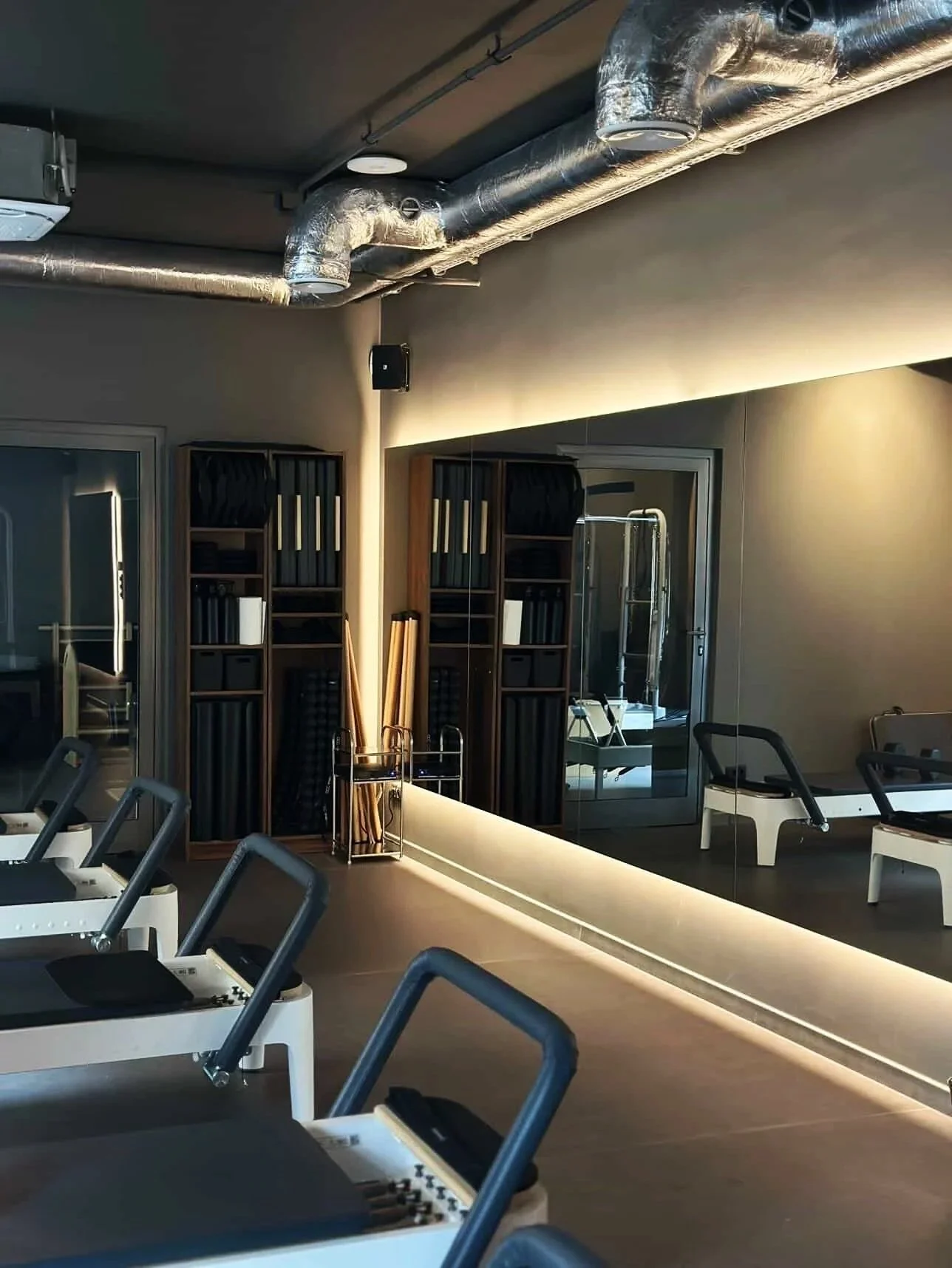 Indoor fitness studio with exercise machines, mirrors, shelving with workout gear, and a mirrored wall.