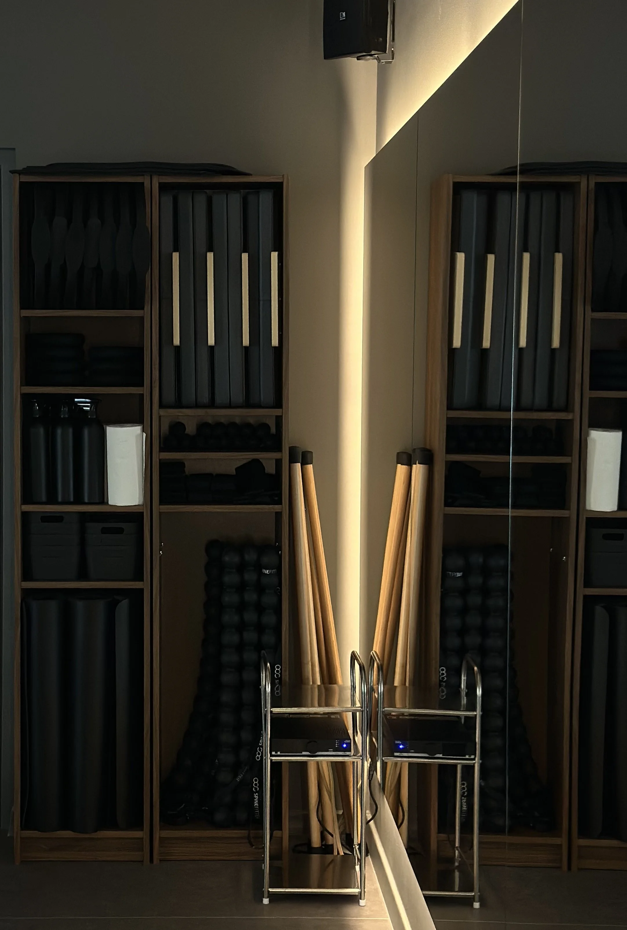 Pilates storage with wooden shelves holding black and gray sport equipment and two wooden rolling pins, alongside a metal cart and a mirror reflecting the shelves, with a wall-mounted radio and speakers