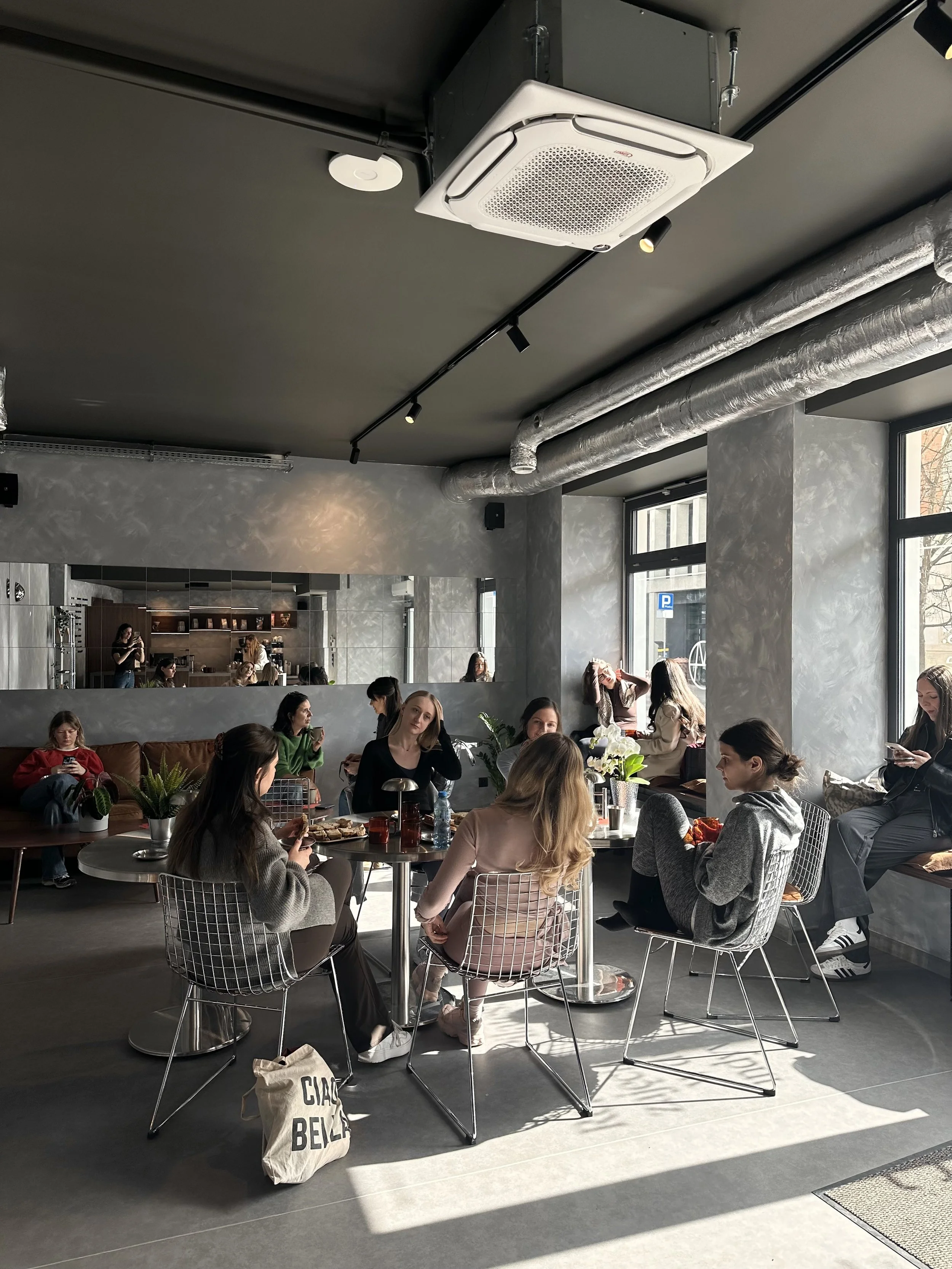 A group of people sitting at tables inside a modern cafe or restaurant, with large windows letting in natural light. Some people are engaged with their phones or talking, while others are enjoying food and drinks. The interior features exposed piping and sleek, contemporary furniture.