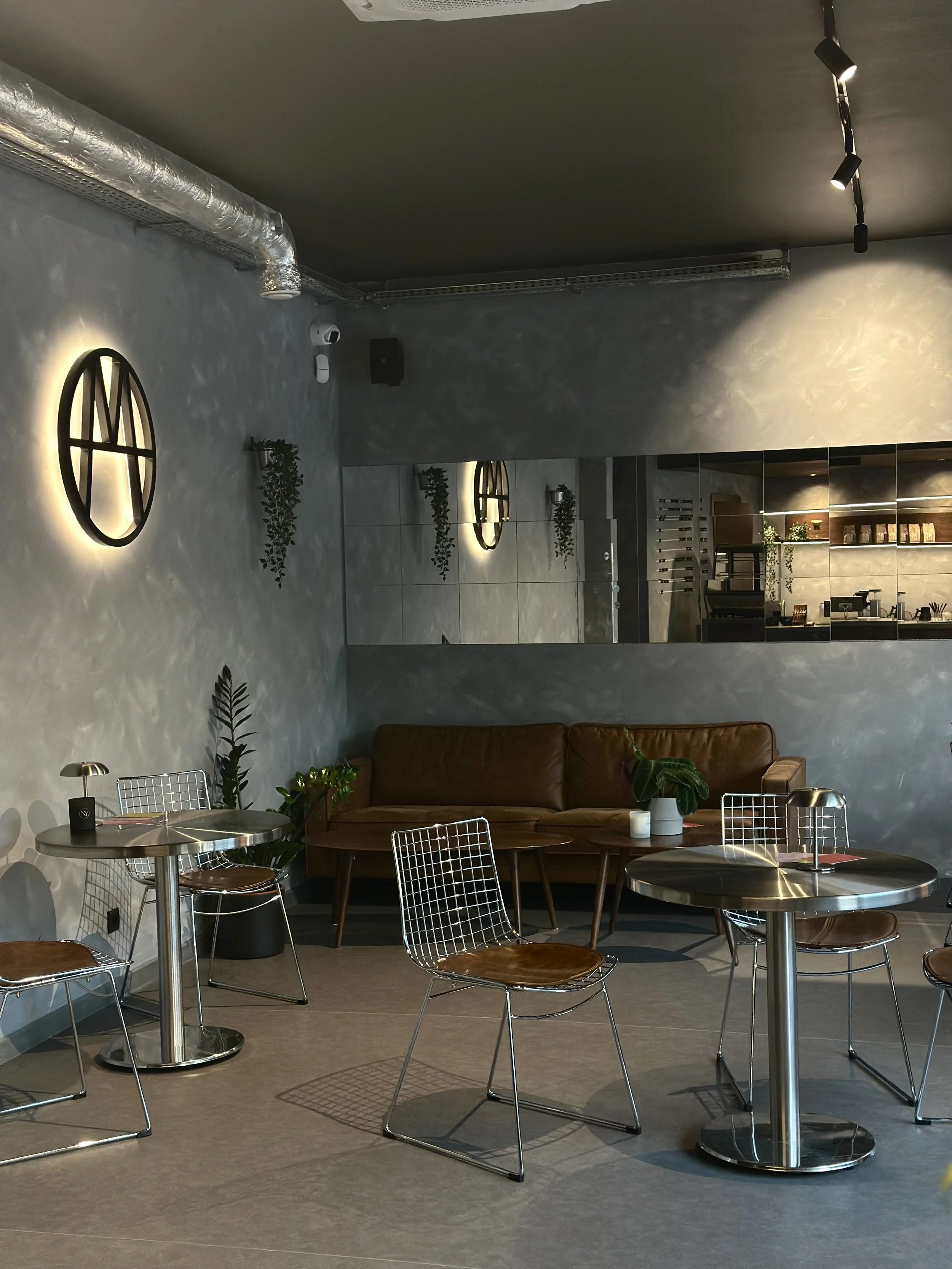The interior of a modern cafe with metal tables and chairs, a leather sofa, decorative plants, wall lighting, a kitchen visible in the background, and a parking lot outside.