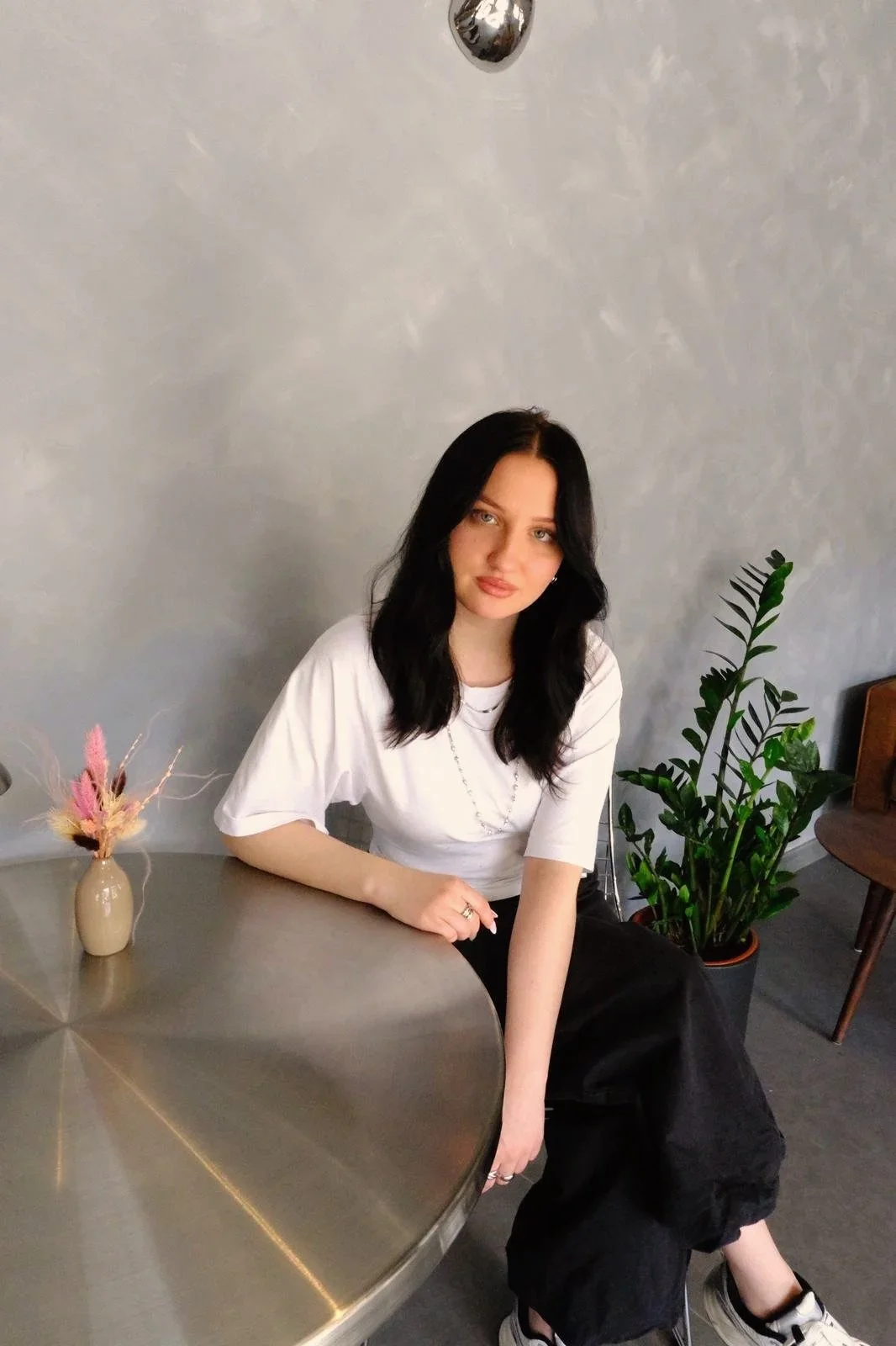 A young woman with black hair and light skin sitting at a round metallic table in a modern room. She is wearing a white t-shirt and black pants, with a silver ring on her finger. There is a beige vase with dried decorative flowers on the table. Behind her, there is a large green plant and a wooden chair. The wall in the background is light gray.