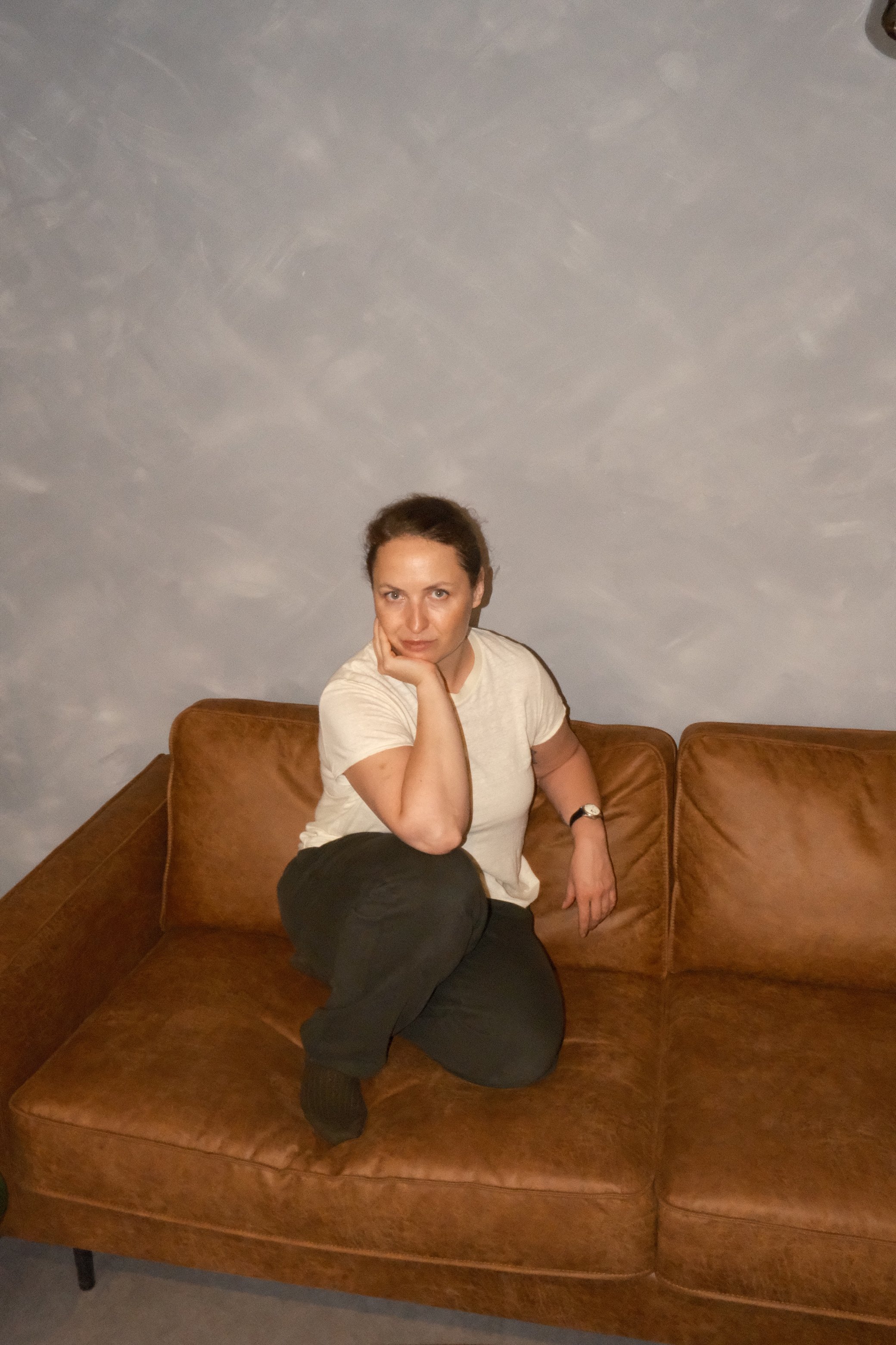 A woman is sitting on a brown leather couch with one leg crossed over the other, resting her head on her hand, in a room with a textured grey wall.