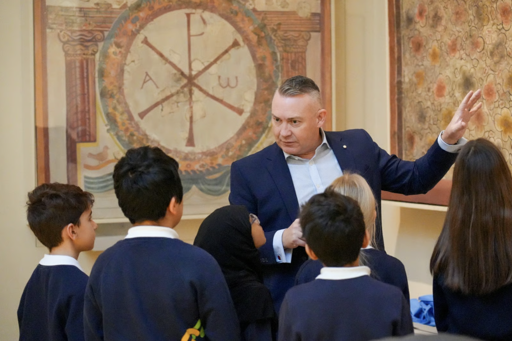 A man in a navy suit and light shirt is talking to a group of students in uniforms inside a room with decorated walls and a large wall clock painting in the background.