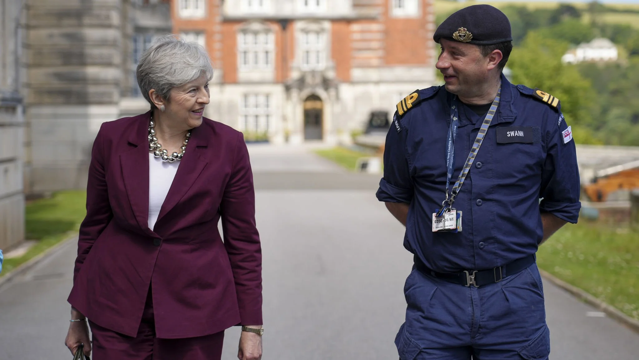 Former Prime Minister Theresa May walking with Royal Navy officer with a cap, badges, and an ID badge, in front of Royal Navy Academy.
