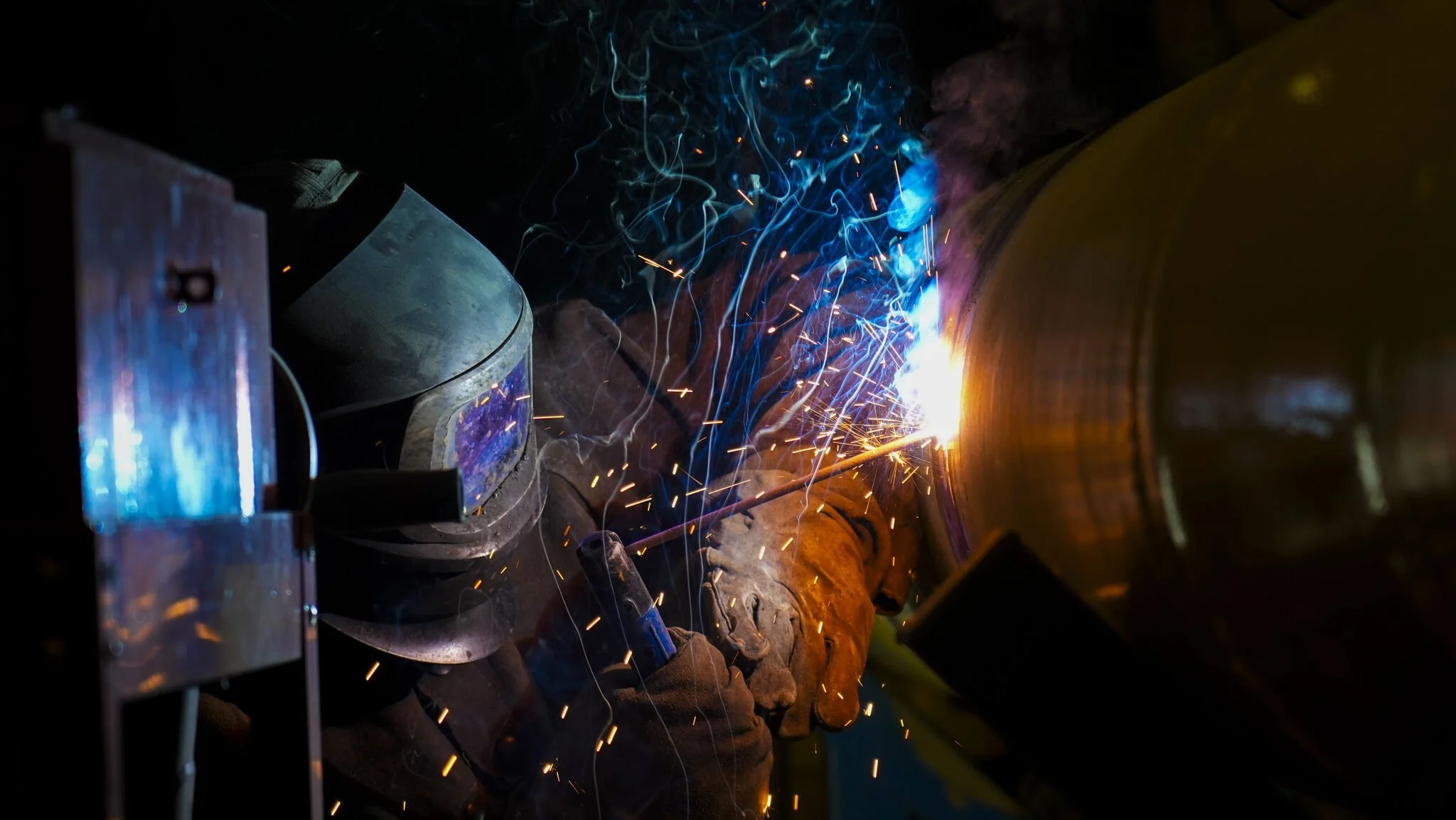 A welder working with a metal pipe, producing bright sparks and blue light from welding arc.