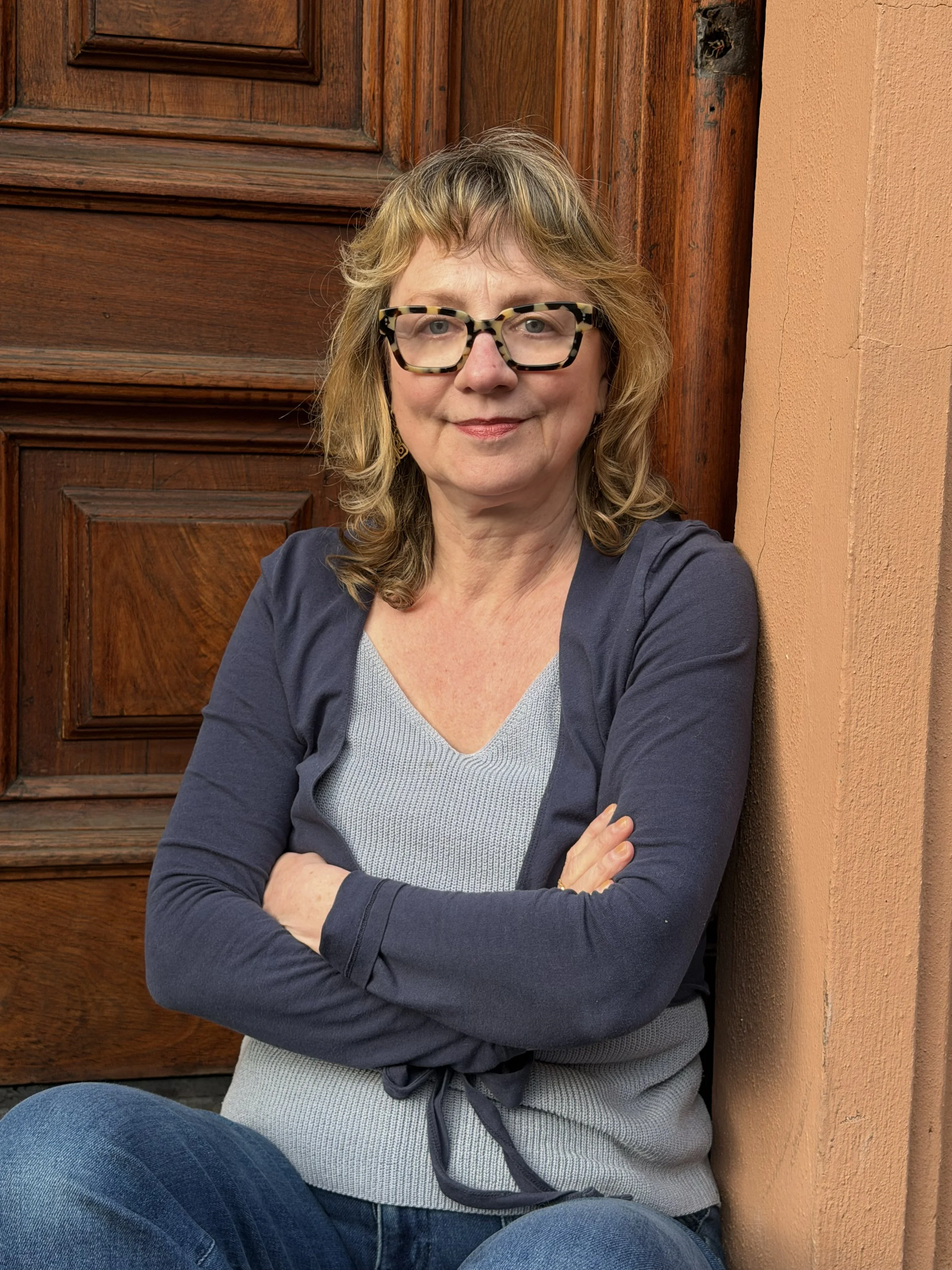 A woman with shoulder-length blonde hair wearing glasses, a gray sweater, and a navy blue jacket, sitting on the floor with arms crossed in front of a wooden door and a peach-colored wall.