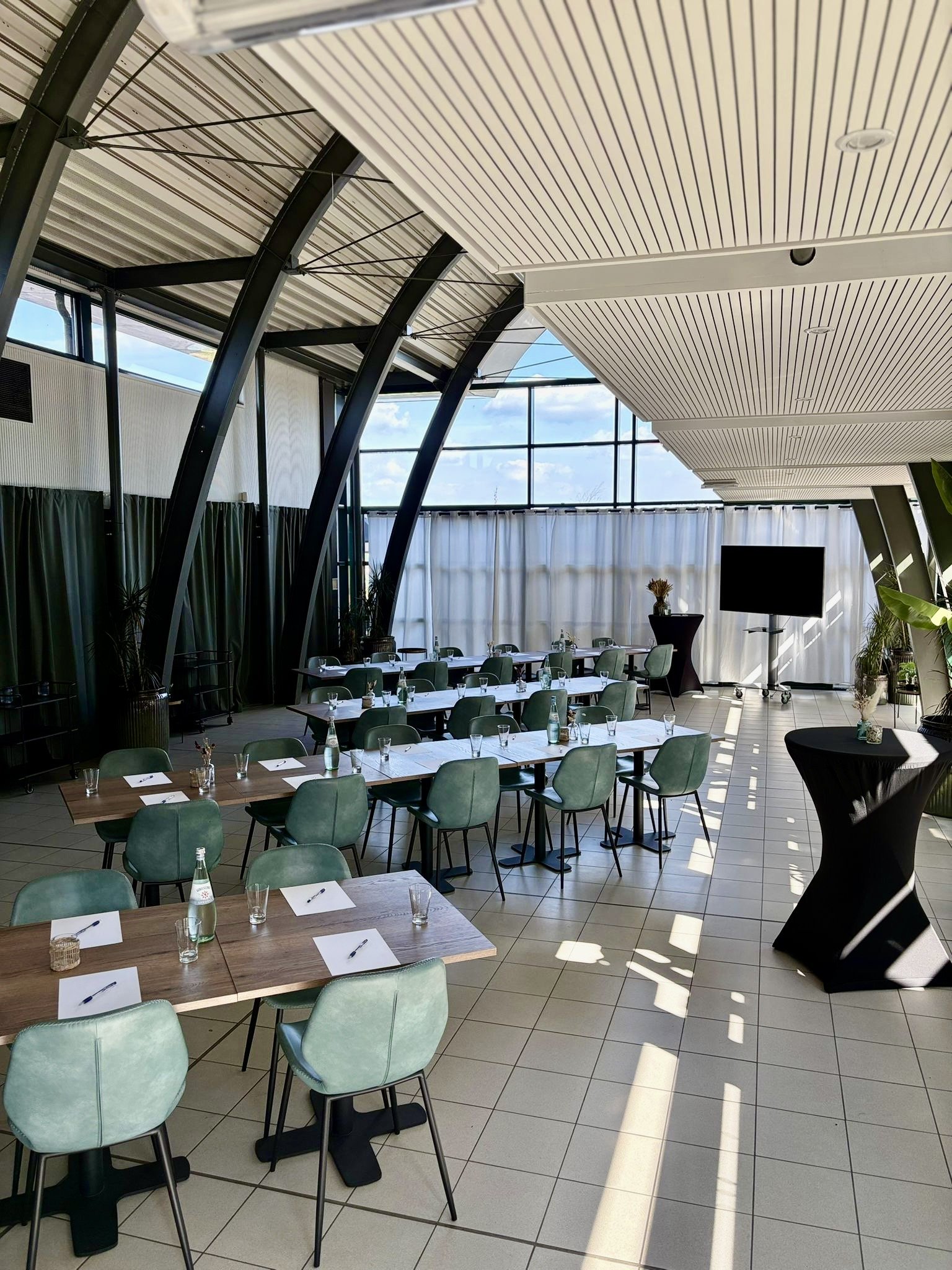 Vergaderruimte met tafels en stoelen opgesteld voor een meeting, grote ramen, een tv en planten.