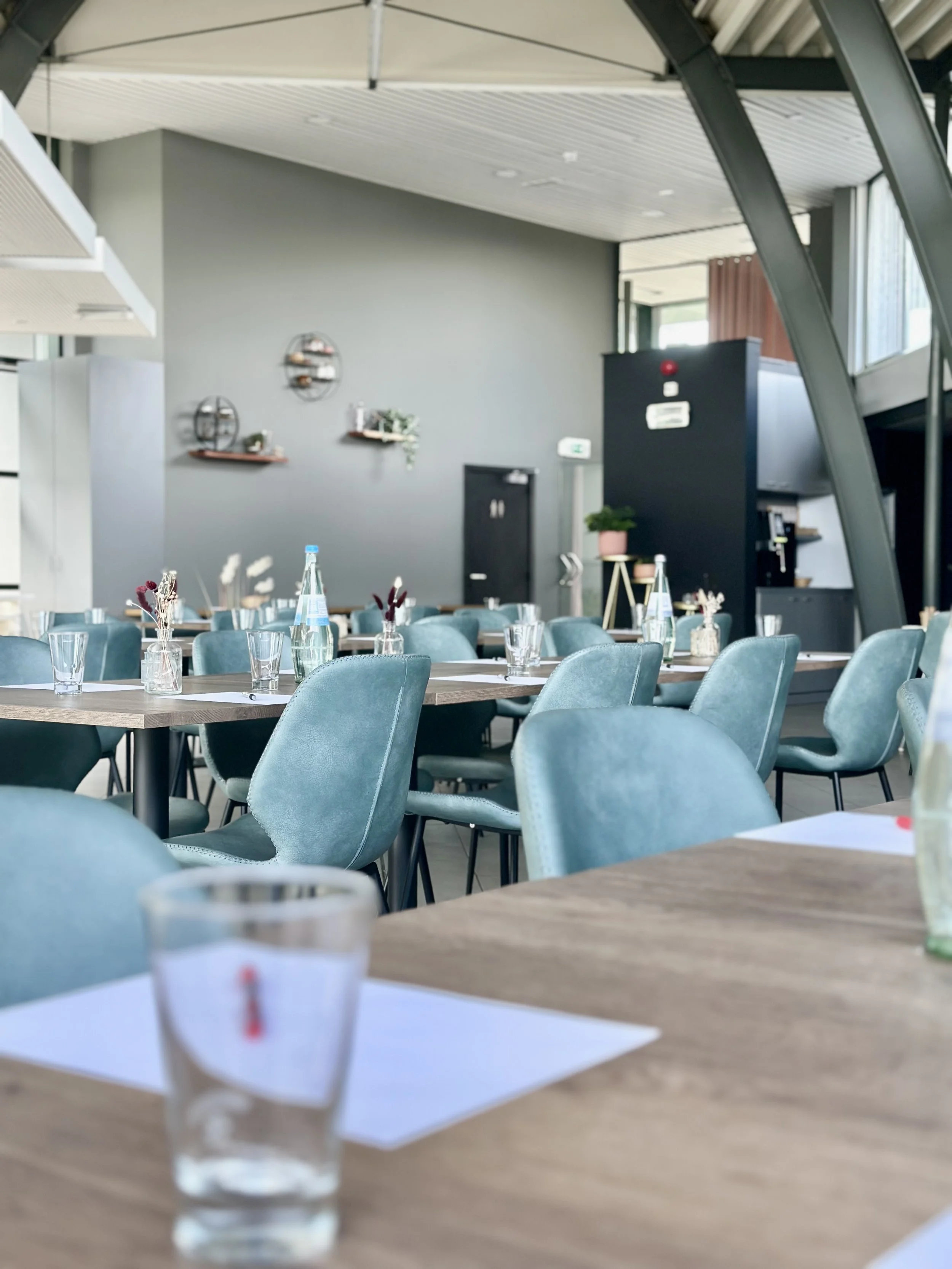 Lege eetruimte met blauwe stoelen, glazen flessen en glazen op de tafels in een modern restaurant, met een grijze muur en decoratieve wandplanken op de achtergrond.