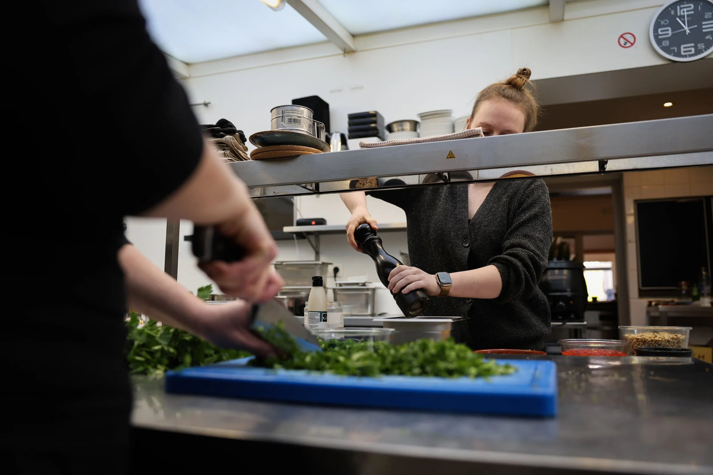 Twee personen die eten bereiden in een keuken: één snijdt kruiden op een blauwe snijplank, de ander giet olie of azijn.