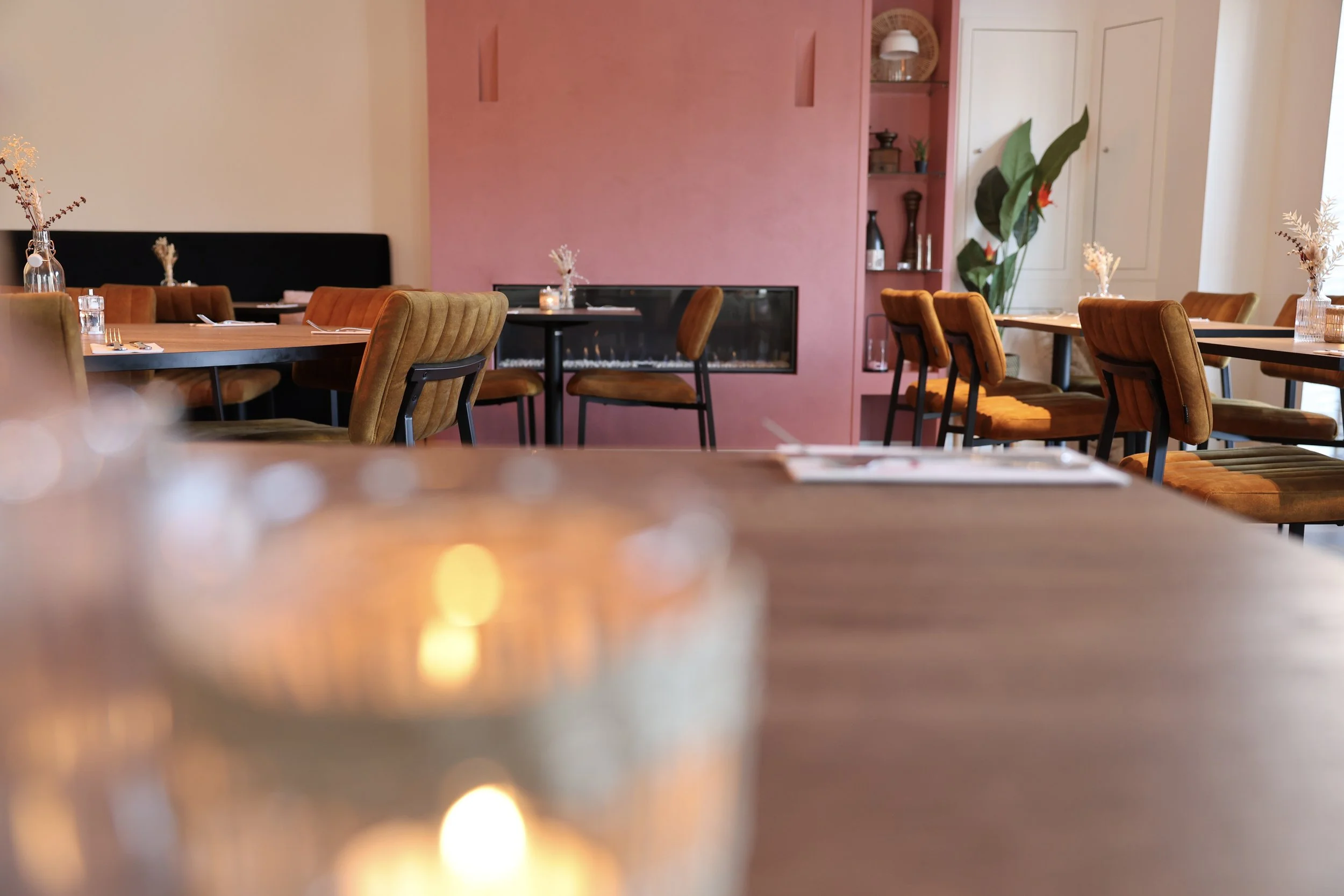 Restaurantinterieur met houten tafels, bruine gestoffeerde stoelen, decoratieve vazen met bloemen, een roze accentmuur met ingebouwde rekken en een grote groene plant bij witte deuren.