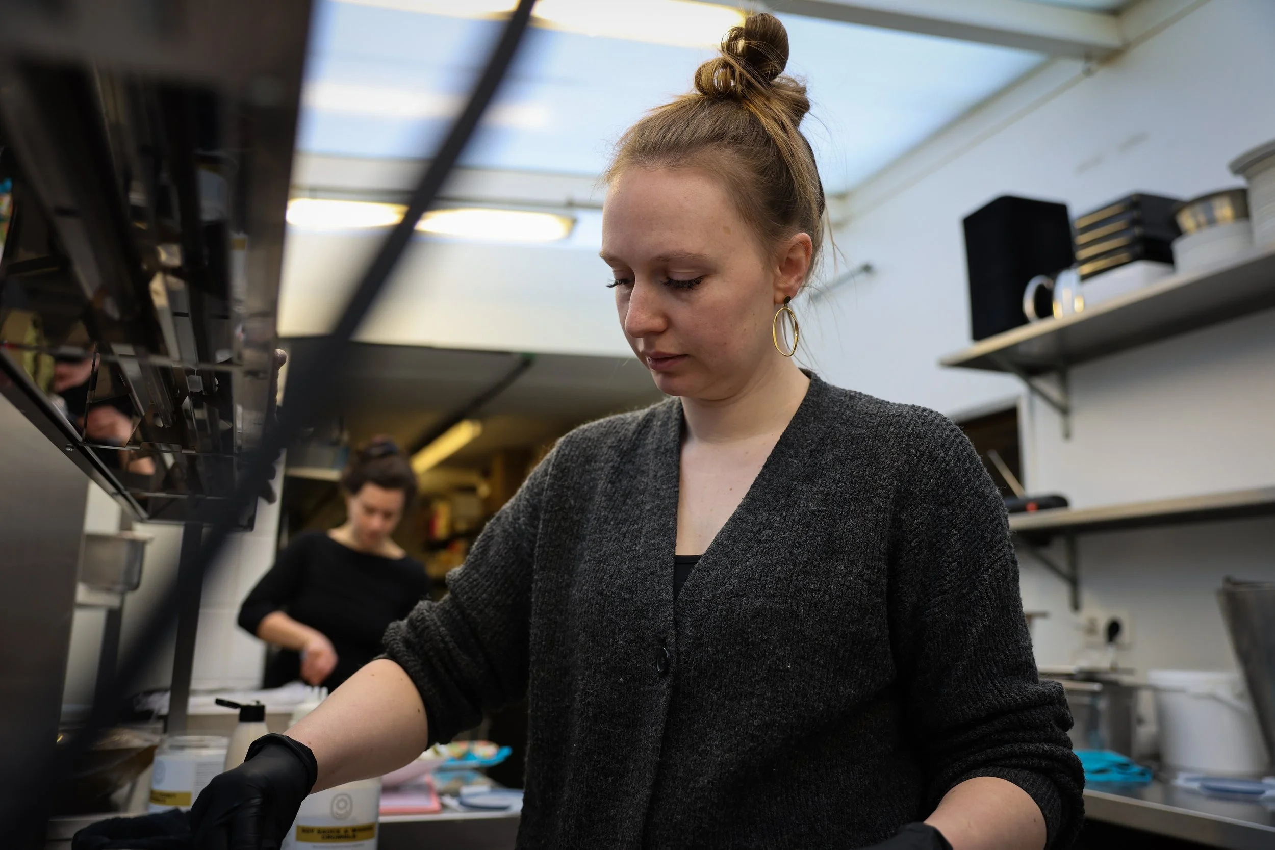 Jonge vrouw met blonde knot, grijze trui en grote oorringen die werkt in een keuken of labo, met een collega op de achtergrond en wandrekken vol materialen.