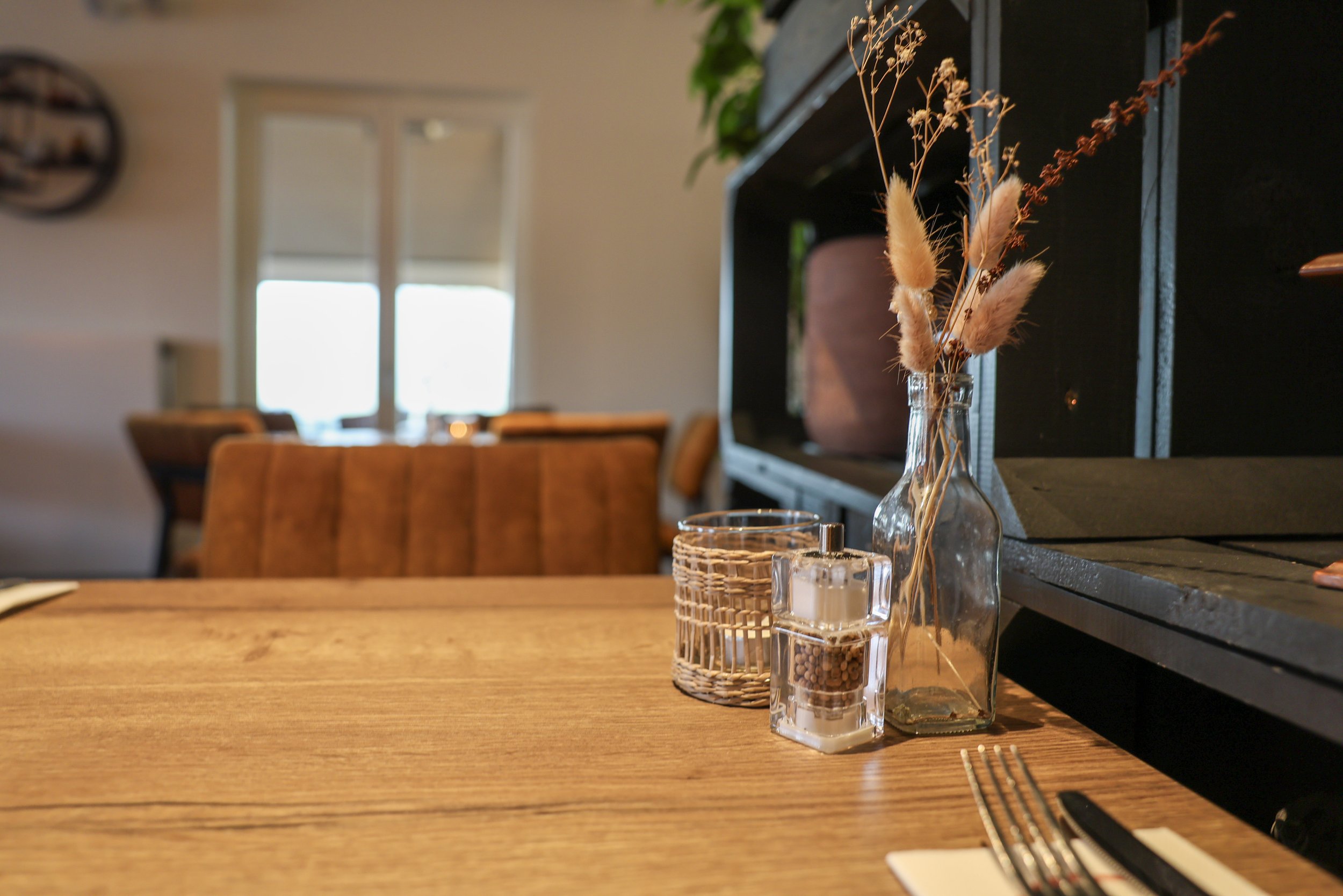 Houten eettafel gedekt met bestek, servet, glazen peperkorrels, een gevlochten mandje en een vaas met gedroogde roze en beige bloemen, in een warm restaurantinterieur met zachte achtergrondvervaging.