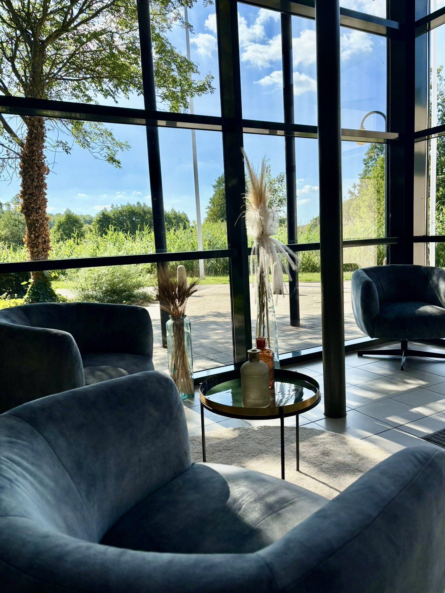 Interieur van een moderne lounge met grote ramen van vloer tot plafond die uitkijken op een groen landschap met bomen en gras onder een blauwe lucht met wolken.