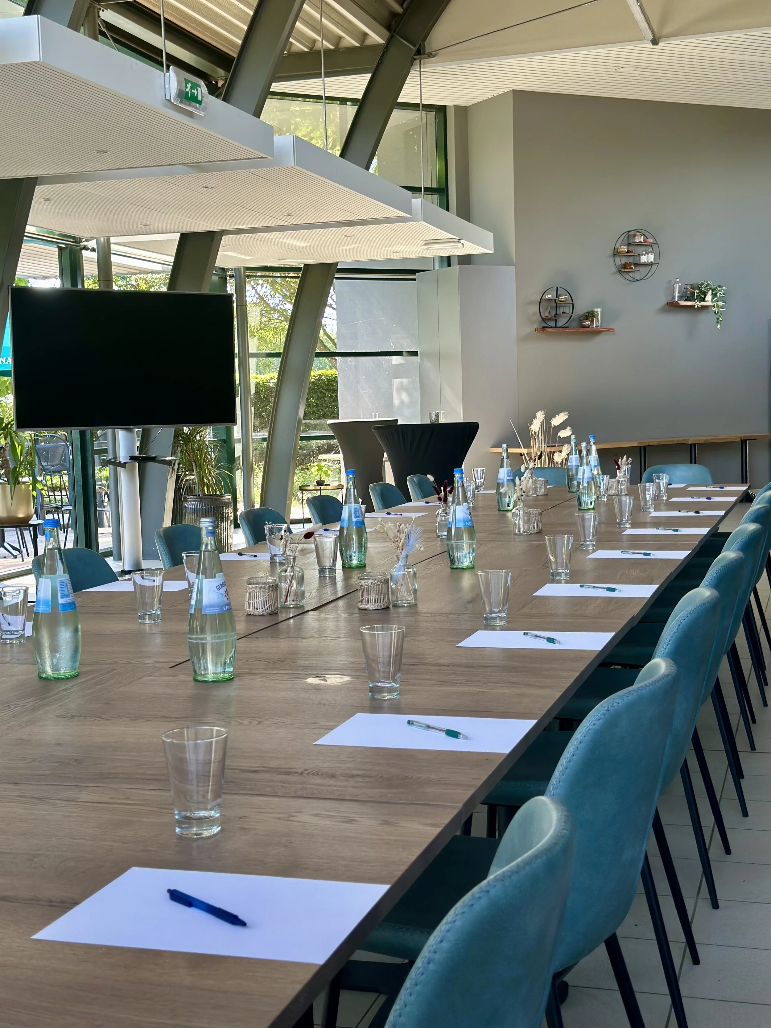 Vergaderruimte met een lange houten tafel met blanco papieren, pennen, waterflessen en glazen. De ruimte heeft grote ramen, moderne inrichting en blauwe gestoffeerde stoelen.