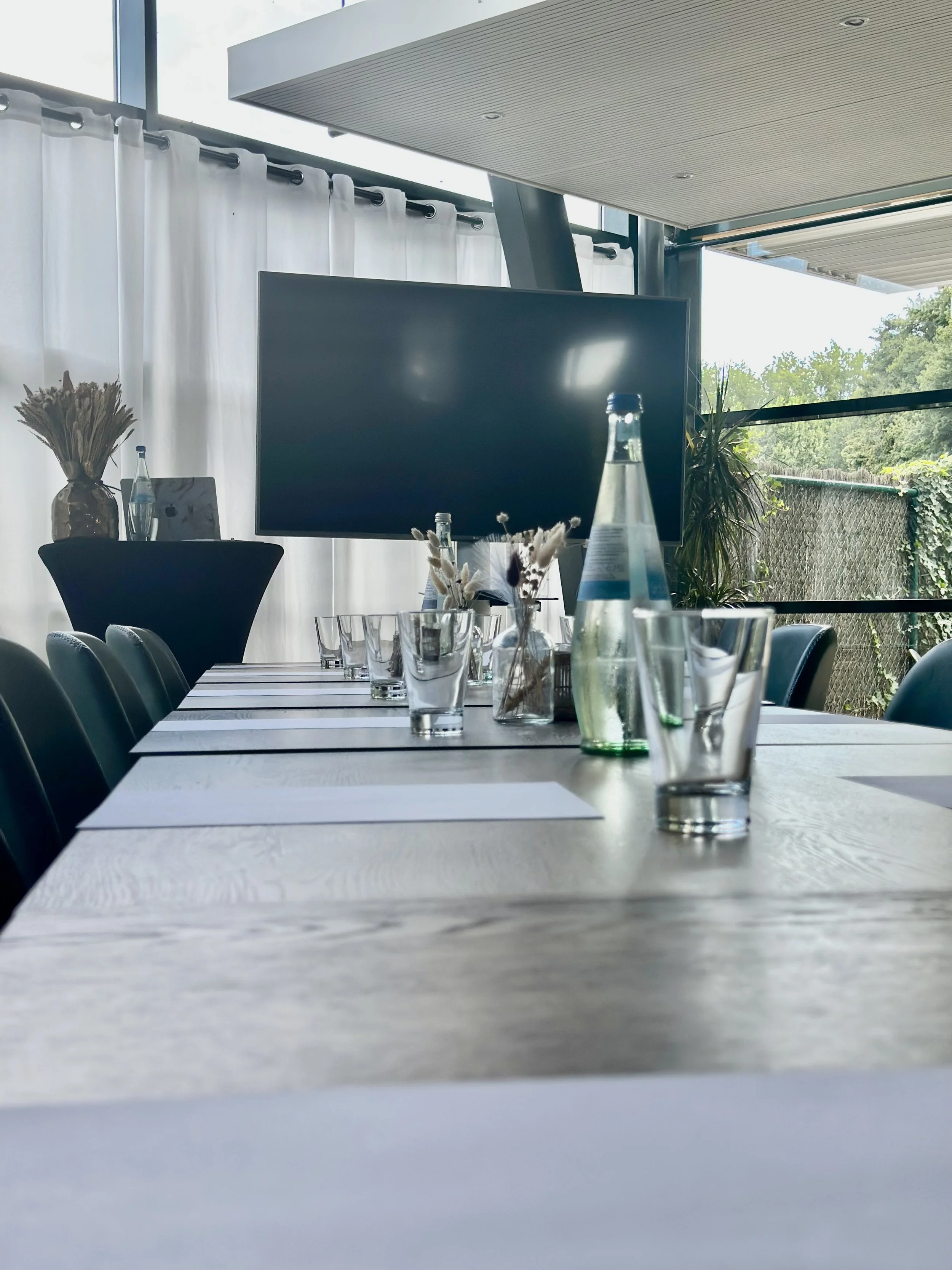 Vergaderruimte met een lange tafel gedekt met glazen, waterflessen en decoratieve droogbloemen, voor een groot flatscreen en ramen met witte gordijnen en uitzicht op groen.
