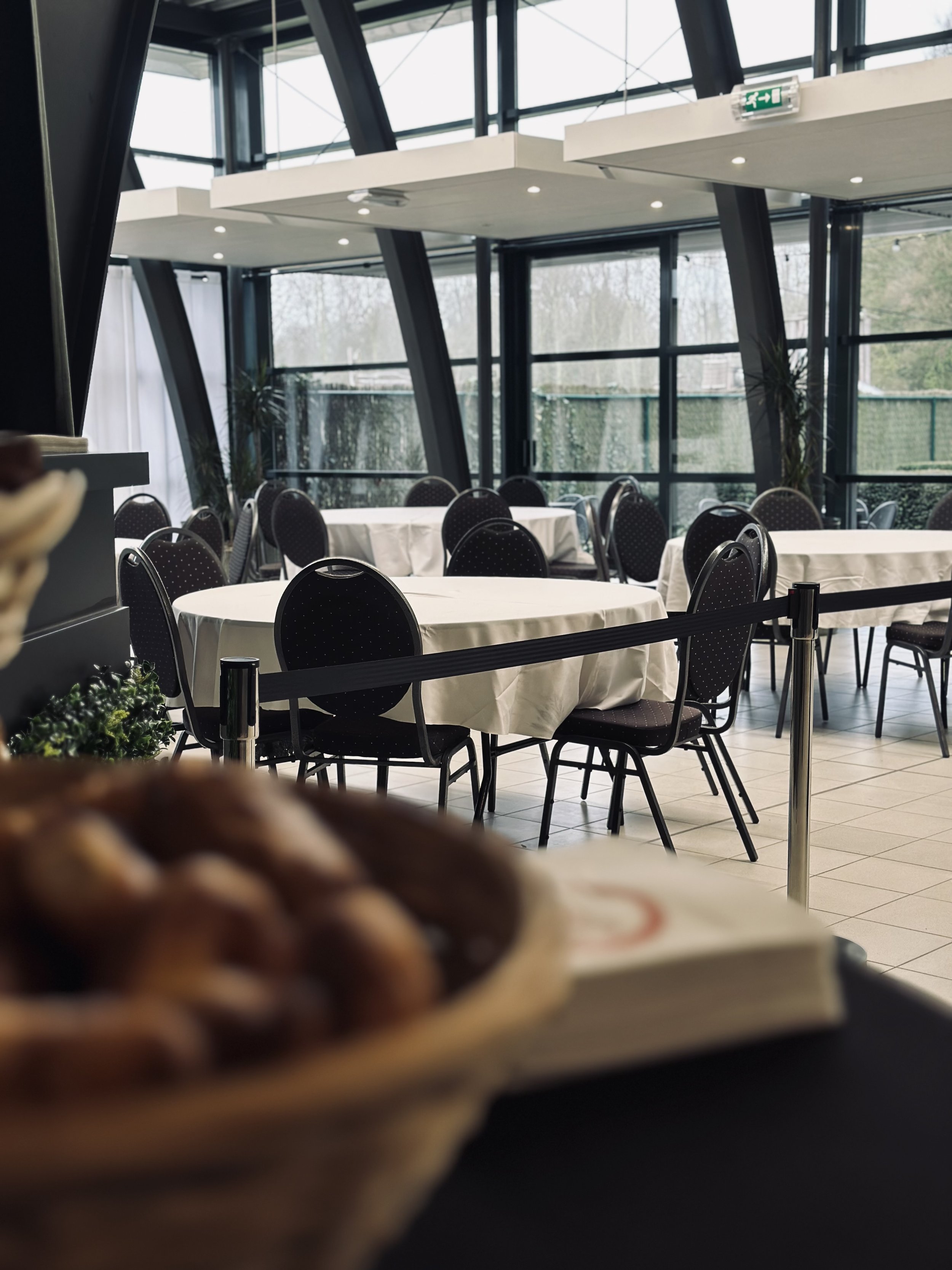 Leeg restaurant met ronde tafels met witte tafellakens en zwarte stoelen, grote ramen met uitzicht op bomen buiten en een schaal met gebak op de voorgrond.