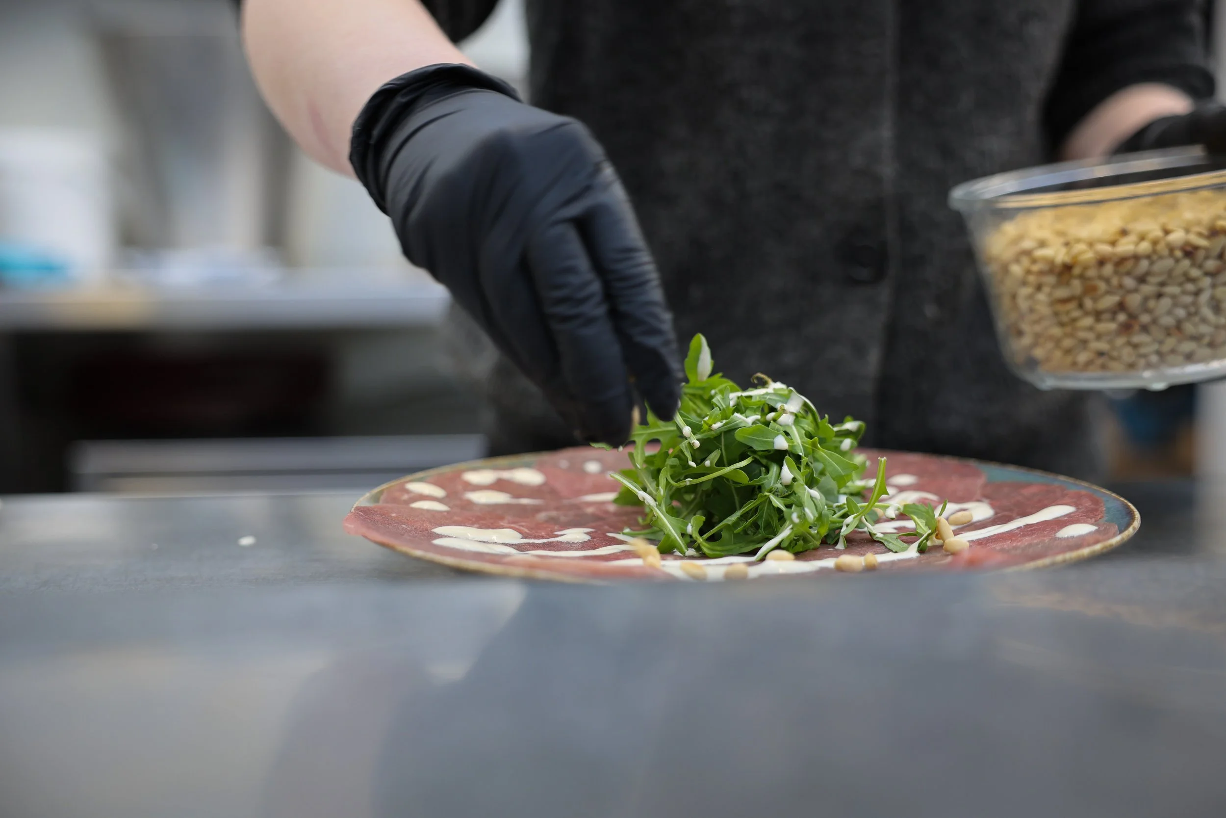 Kok met zwarte handschoenen die een gerecht bereidt met dungesneden vlees, microgroenten, witte saus en pijnboompitten.