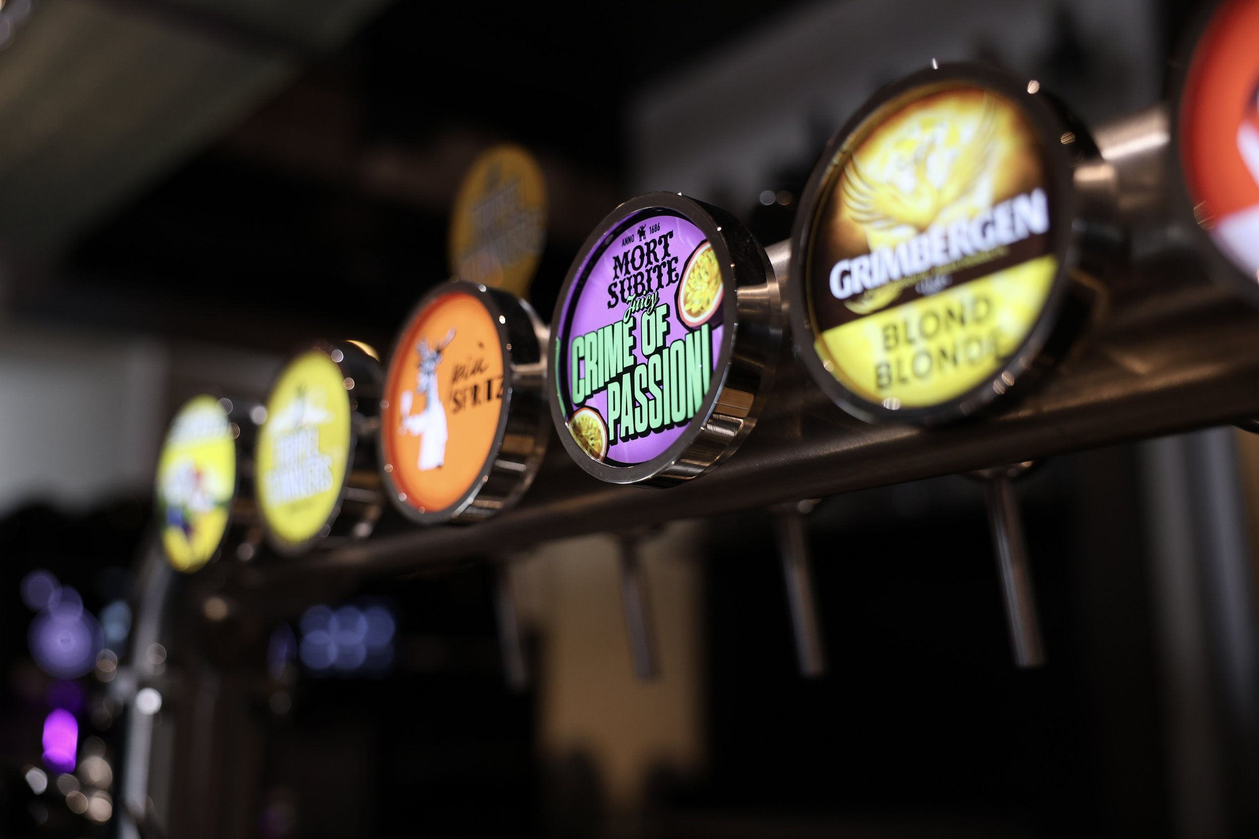 Close‑up van kleurrijke biertapkraantjes met verschillende brouwerijlogo’s en biernamen.