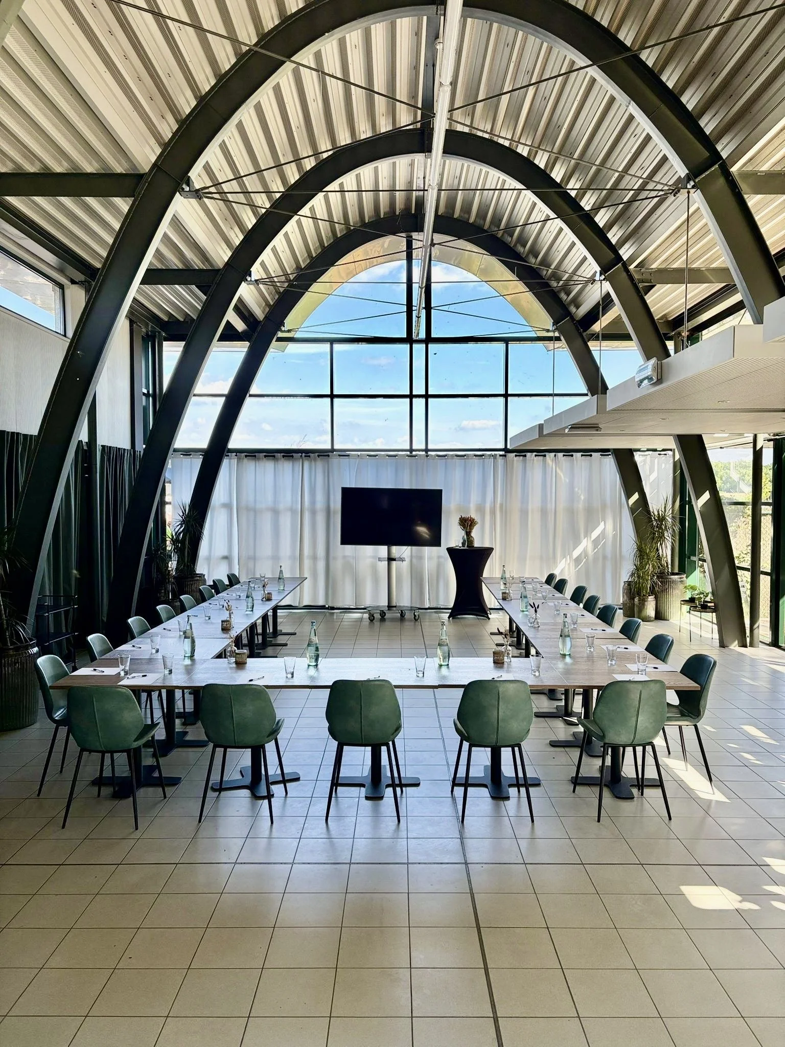 Vergaderruimte met een U-vormige tafelopstelling, groene stoelen, waterflessen en glazen, en een groot flatscreen op een standaard, in een modern gebouw met grote ramen en zichtbare stalen boogbalken.