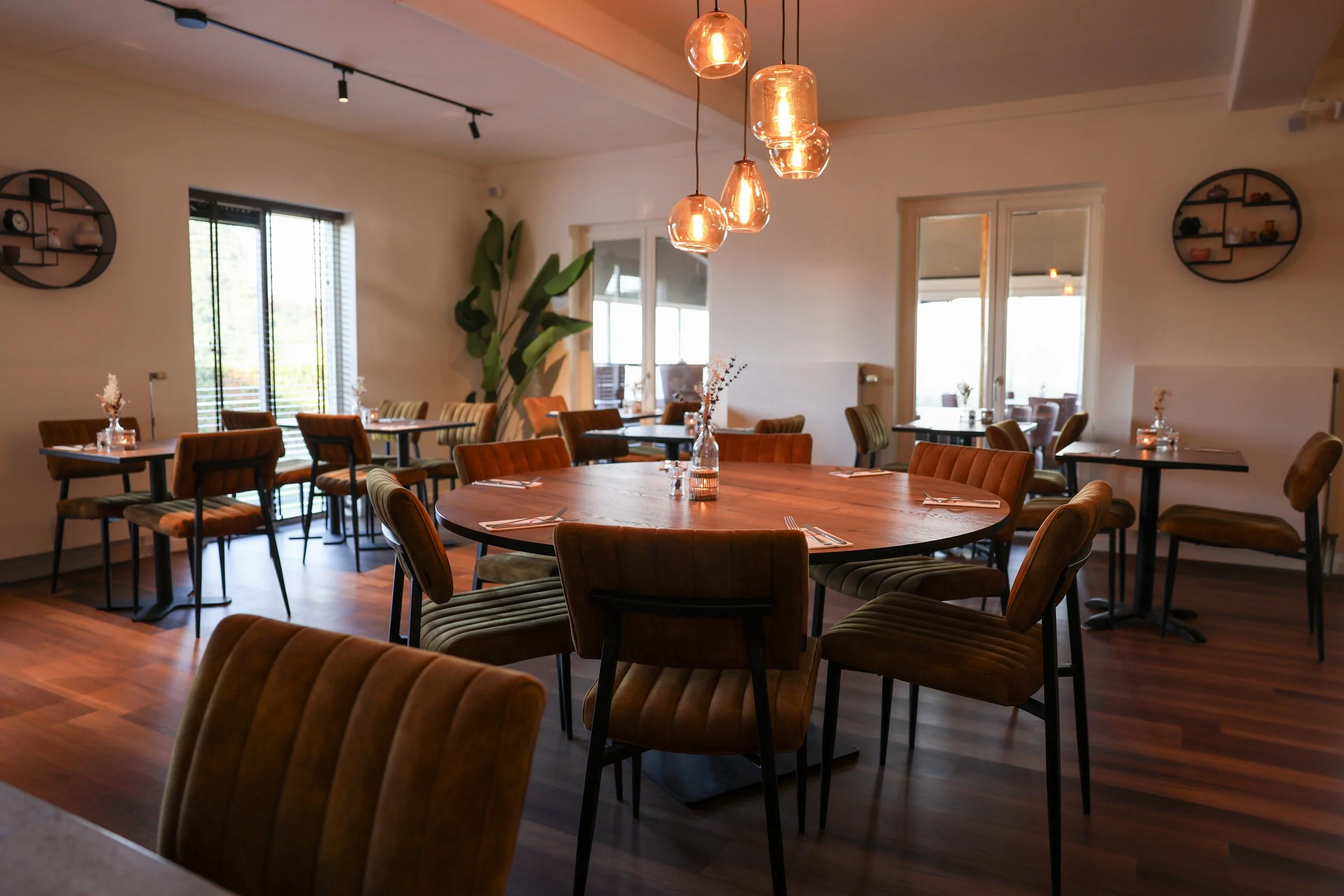 Restaurant met houten tafels, fluwelen stoelen, sfeerverlichting en grote ramen.
