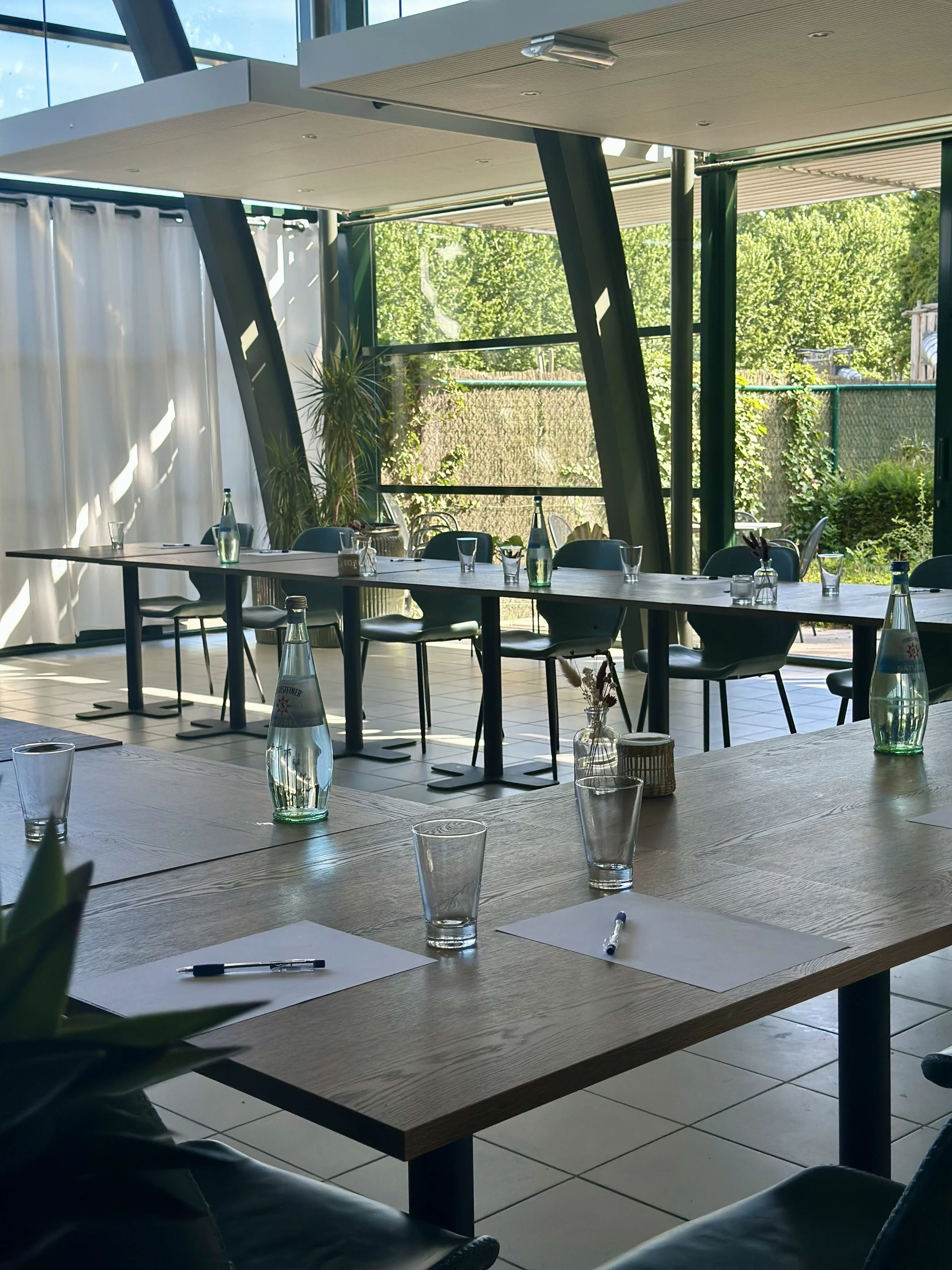 Lege vergader- of conferentieruimte met tafels voorzien van waterflessen, glazen, notitieblokken en pennen, en grote ramen met uitzicht op groene bomen.