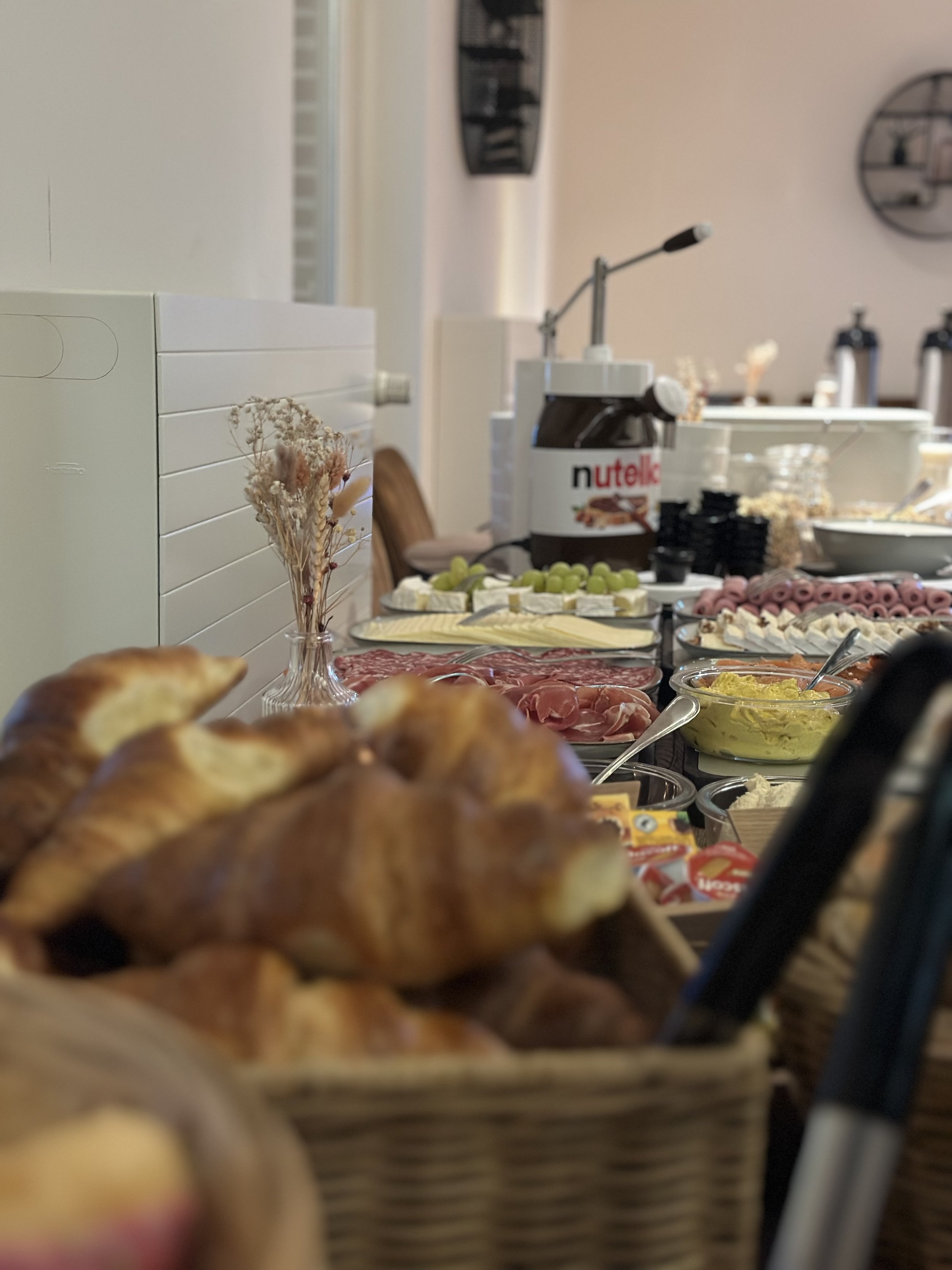 Ontbijttafel met diverse gerechten zoals croissants, fijne vleeswaren, kazen, druiven en smeersels, met op de achtergrond een koffiemachine en keukenaccessoires.