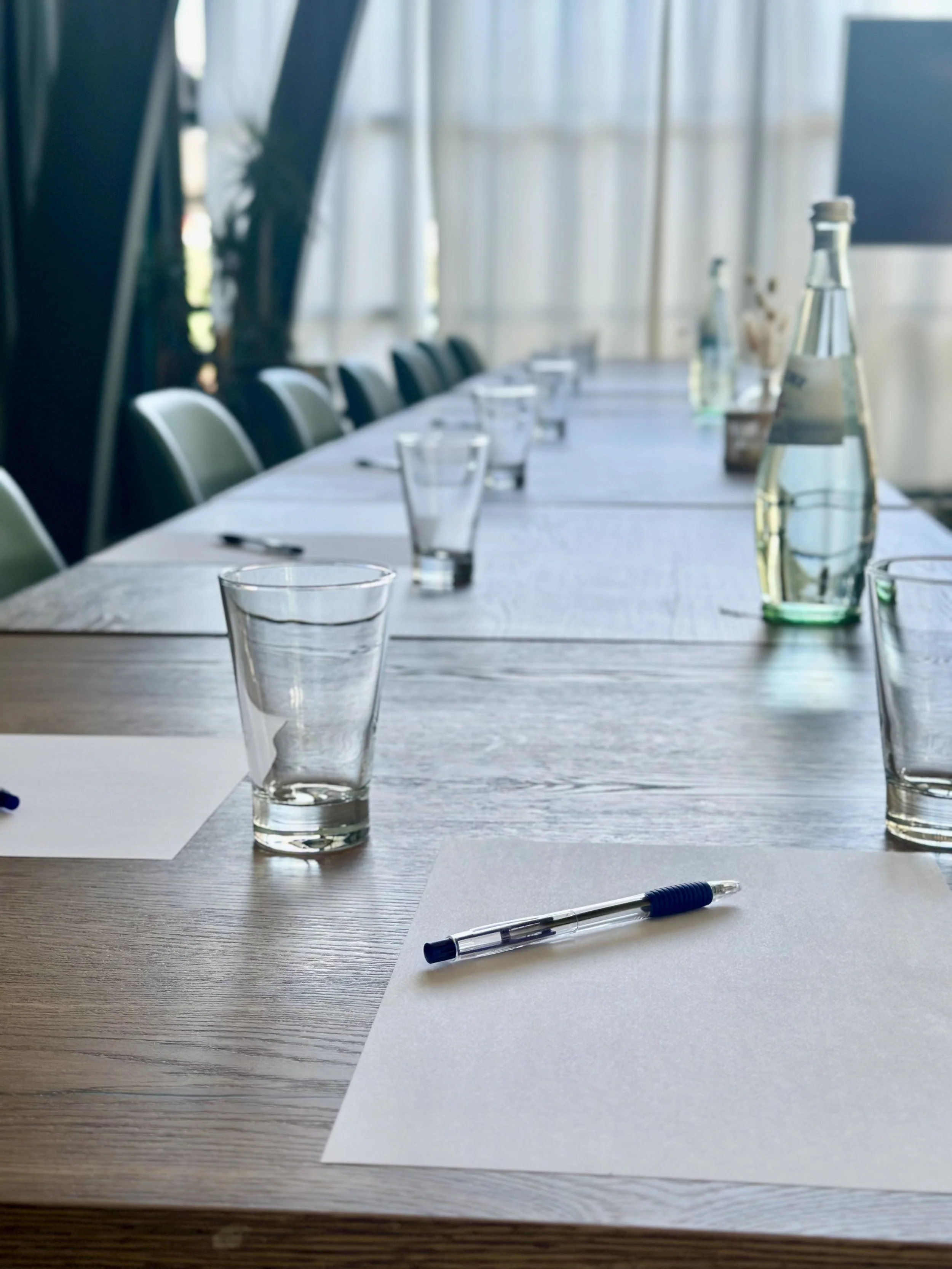 Vergaderruimte met een lange houten tafel met lege glazen, waterflessen en blanco notitiebladen met pennen. De ruimte heeft grote ramen met gordijnen en stoelen langs de tafel.