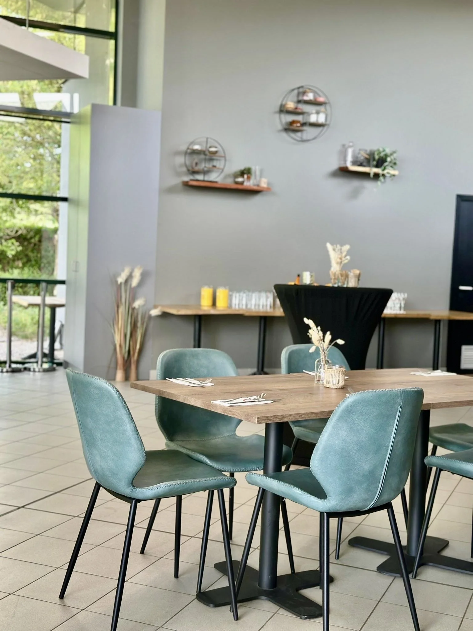 Modern restaurantinterieur met een houten tafel, turquoise stoelen en minimalistische decoratie. Grote ramen met zicht op groen, wandrekken met decorstukken en een tafel gedekt met glazen en gele kaarsen.