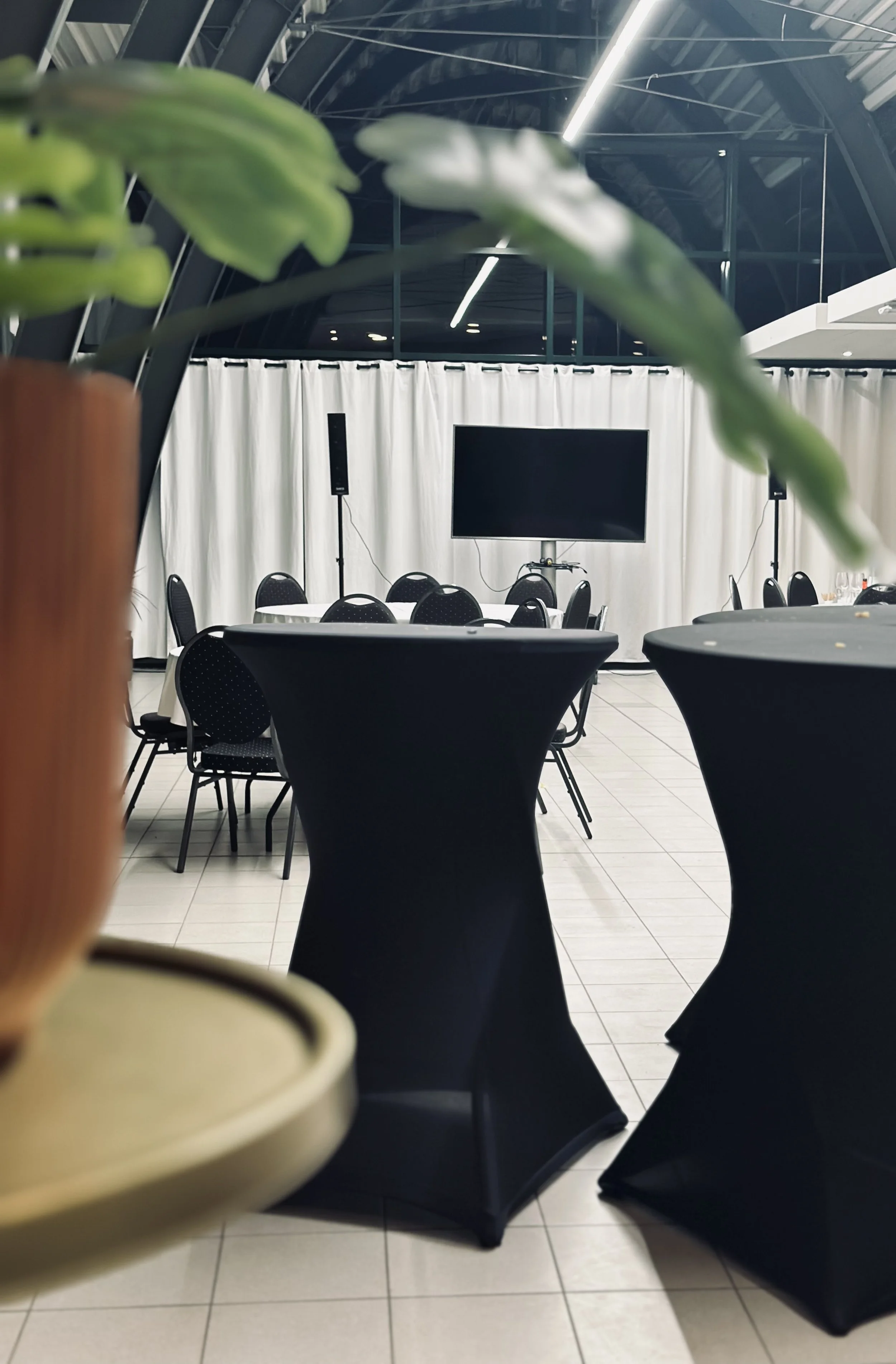 Vergader- of eventruimte met ronde tafels met zwarte en witte tafellakens. Vooraan staat een groot scherm met audioapparatuur, met witte gordijnen als achtergrond.