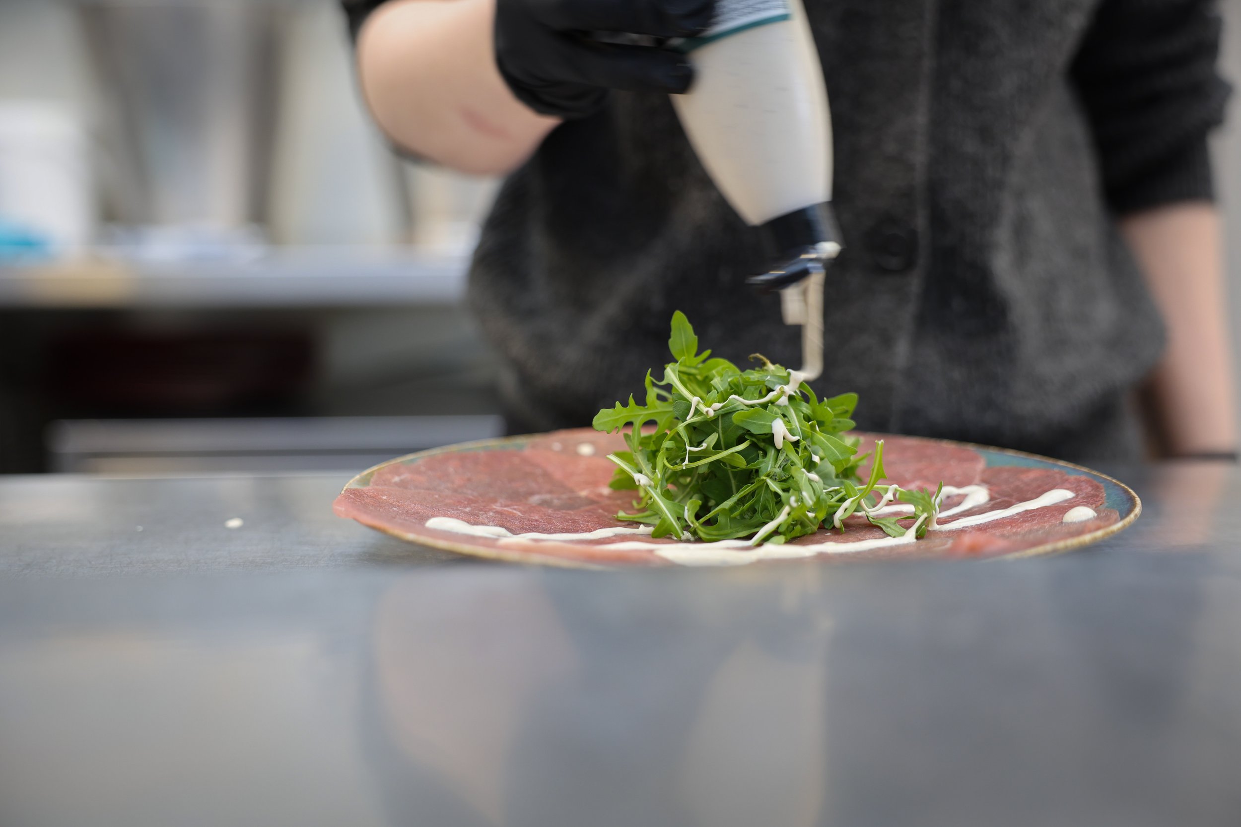 Persoon die verse groene kruiden op een plak rauw vlees legt, afgewerkt met witte saus.