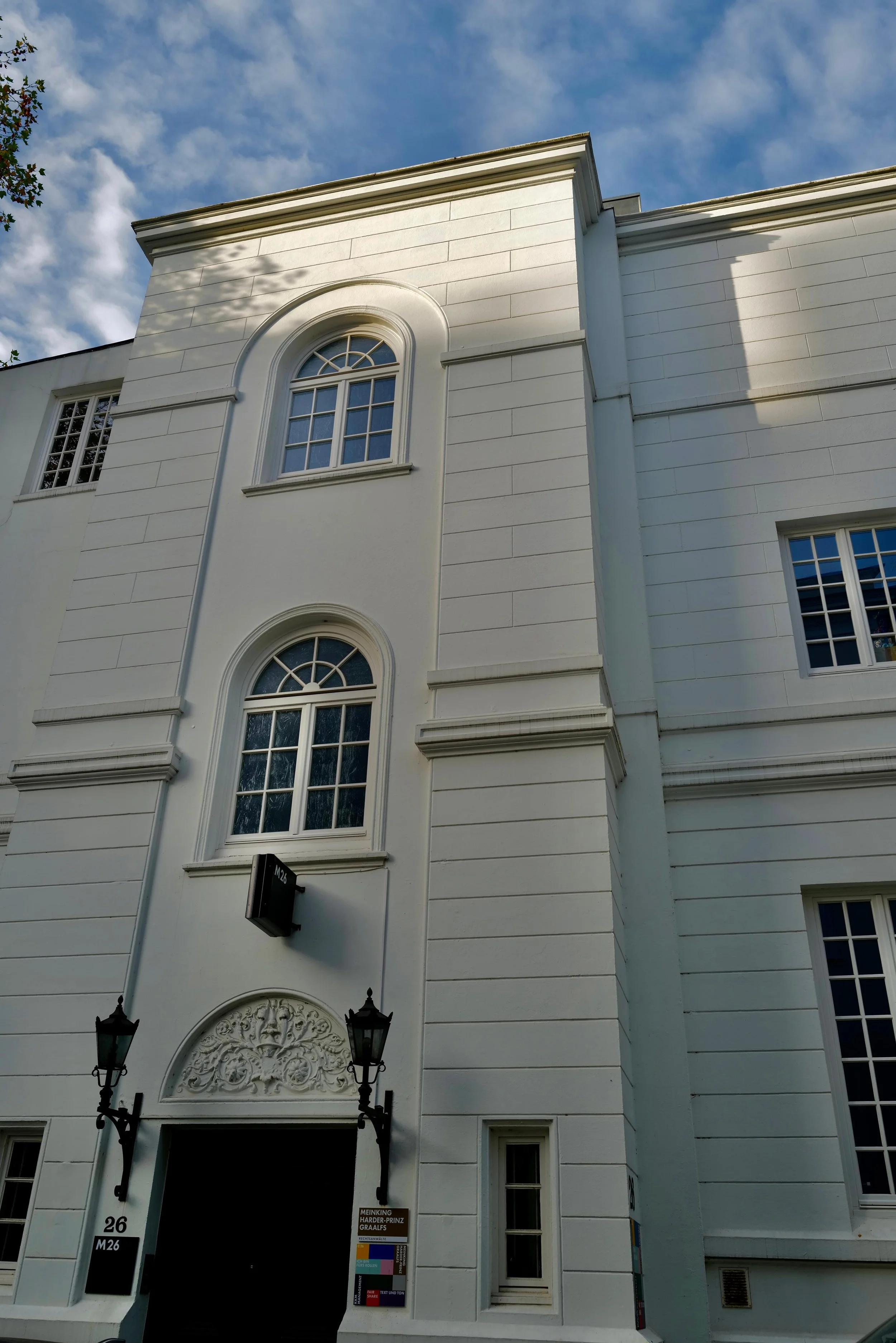 Ein weißes Gebäude mit mehreren fenstern und dekorativen Elementen oberhalb der Tür, zwei Laternen neben dem Eingang, unter blauem Himmel mit Wolken.