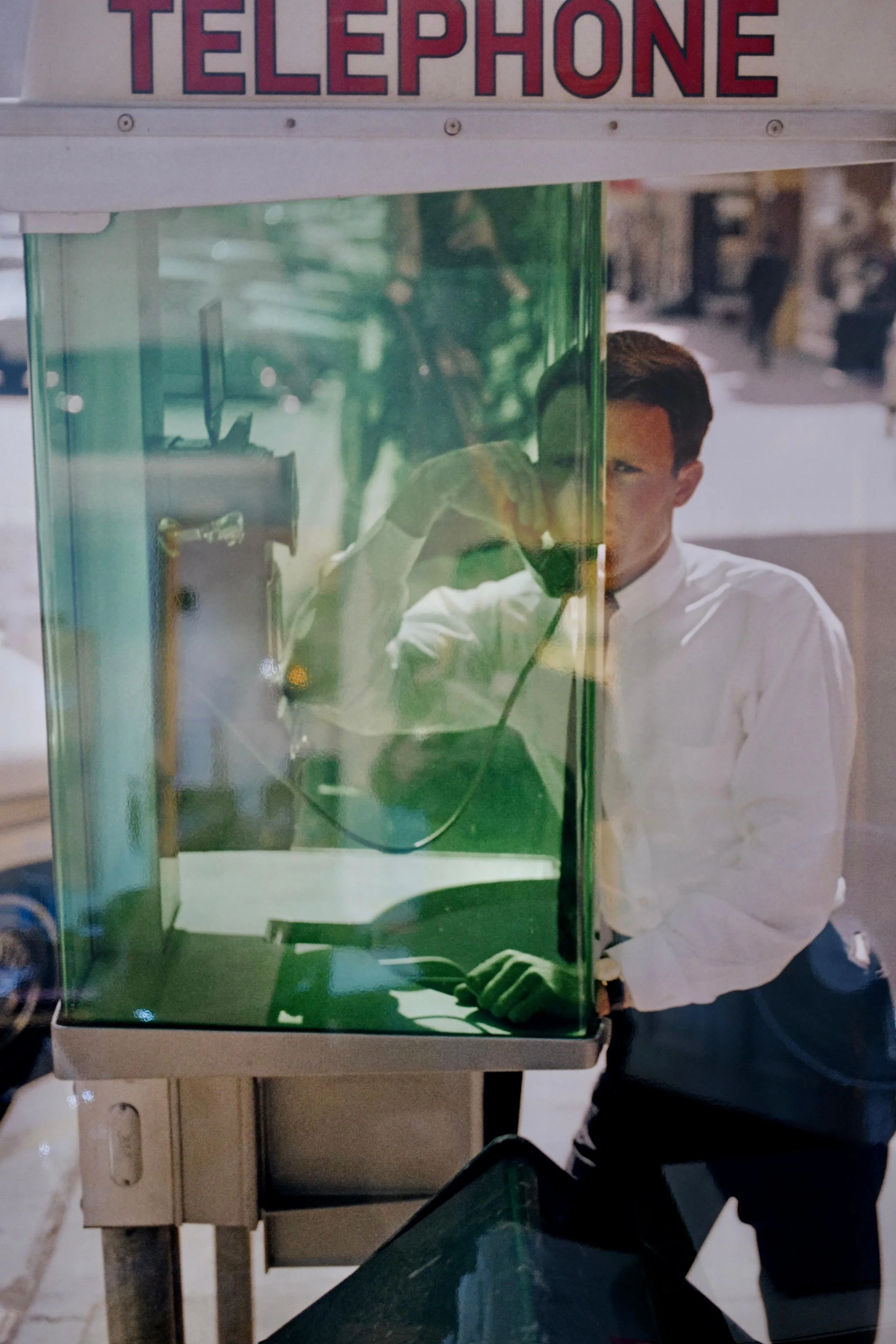Ein Mann in einem weißen Hemd steht an einem öffentlichen Telefonautomaten im Freien und spricht durch das Telefon. Die Aufnahme ist durch die grüne Glaswand des Telefons gemacht, die den Mann teilweise verdeckt und reflektiert. Im Hintergrund sind verschwommene städtische Umgebung und Passanten sichtbar.